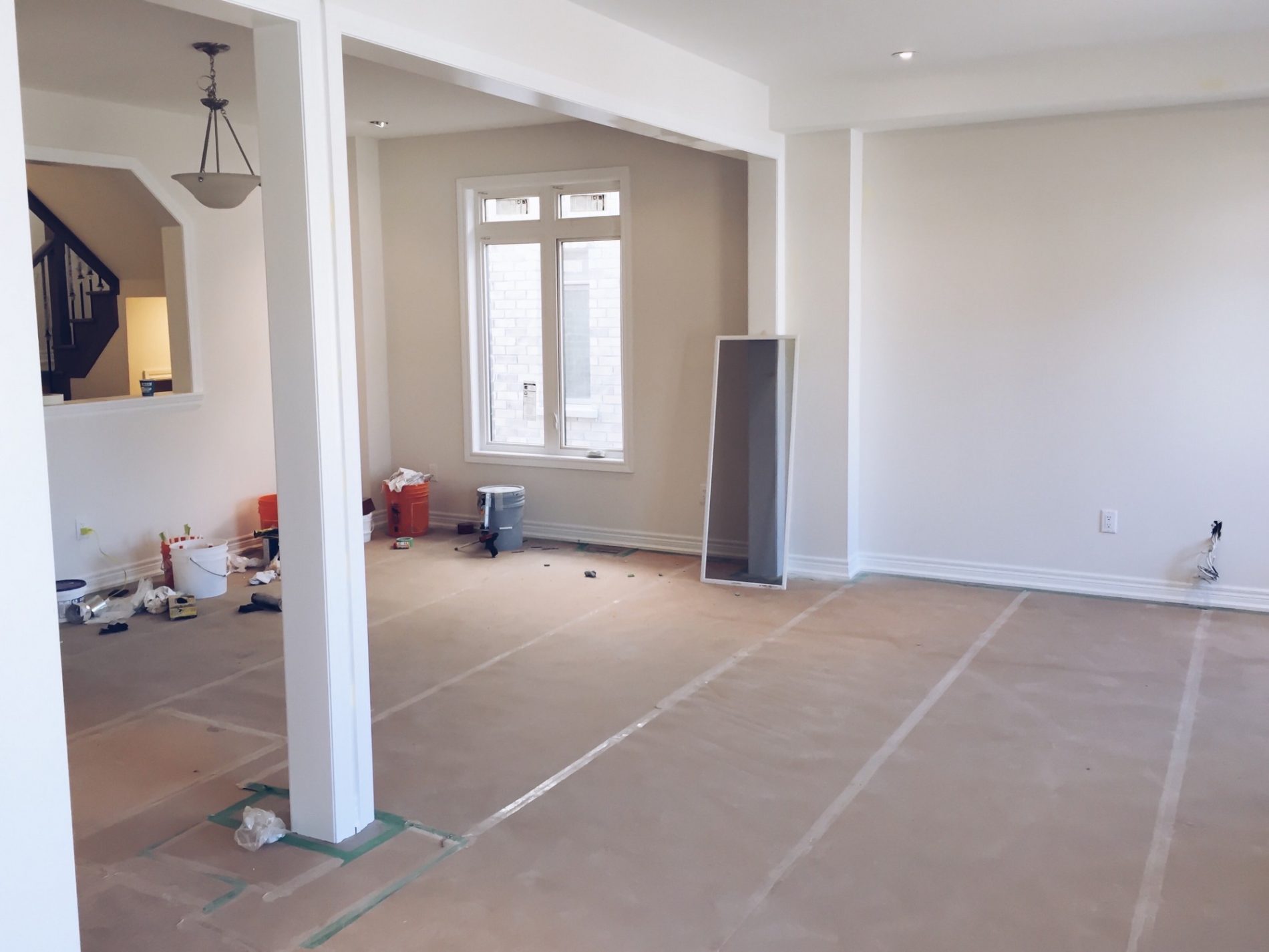 This is an image of an interior space that appears to be under renovation or redecoration. The floor is covered with protective paper, suggesting that painting or other work that could cause spills or stains is being done. There are various items scattered throughout the room, such as paint buckets, a vacuum cleaner, a step ladder, and other tools and materials typically used in home improvement projects.The room has white walls and features such as window frames and baseboards, indicating that it has been recently painted or is about to be painted. There's also a column in the foreground, which adds a structural element to the space, and various electrical outlets visible on the walls, indicating that this is likely a living space. A tall, narrow mirror or possibly a metallic panel of some kind is leaning against one wall. Light fixtures can be seen attached to the ceiling, and there is a small alcove or hallway leading to another room visible on the left side where a pendant light fixture hangs.The room has large windows providing natural light and there's a sense of openness, suggesting it might be a living room or a similar common area in a residential property. The renovations seem to be in progress.