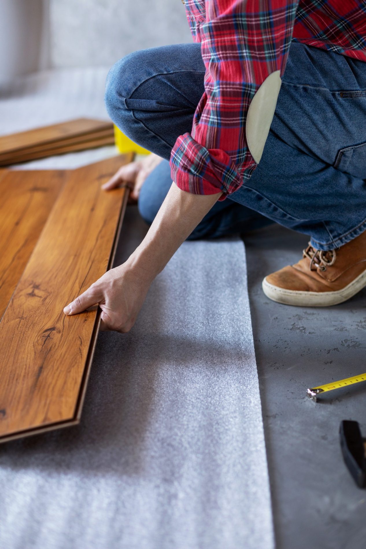 The image shows an individual in the process of installing or laying out laminate flooring. The person is wearing casual attire—blue jeans, a checkered shirt, and brown work boots—and is crouched down pressing a plank of laminate flooring into place. There is underlayment material on the floor beneath the laminate, and a tape measure is also visible on the floor, suggesting that the person has been measuring and cutting the planks to fit. The setting seems to be an indoor space undergoing renovation or decoration.