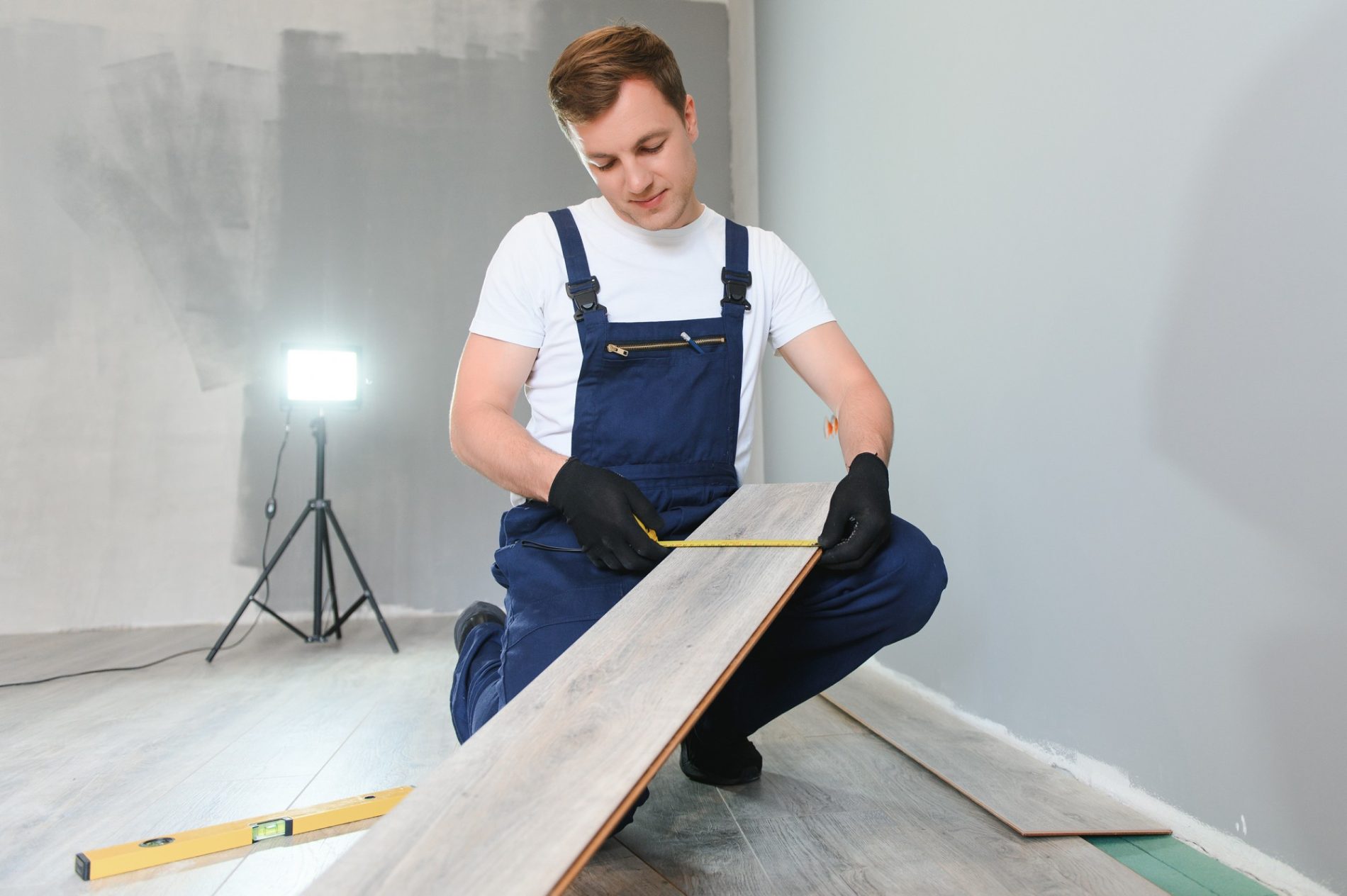 The image shows a man engaging in a flooring installation or repair job. He is measuring a wooden laminate plank with a tape measure, likely preparing to cut it to size to fit the designated area. The man is wearing a white T-shirt under blue overalls and black gloves, which suggests that he's taking precautionary measures to protect his hands. There is also a yellow spirit level on the floor nearby, indicating that precision and levelness are important for this task. The room has a gray wall and the new flooring appears partially installed, with a light and tripod stand visible in the background, which may be used to provide adequate lighting for the work area or to document the process.