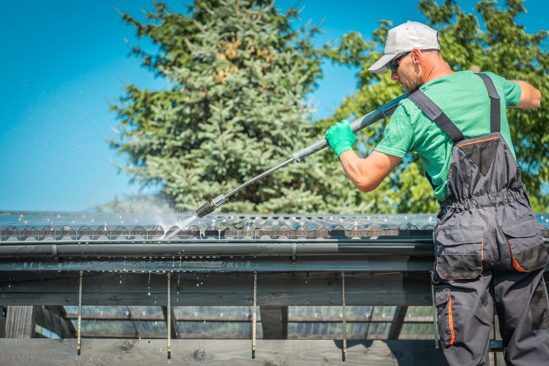 This image shows a person cleaning a solar panel using a high-pressure washer. The individual is wearing a cap, sunglasses, a green t-shirt, protective gloves, and overalls. They are standing on what appears to be a wooden structure, likely constructed for the purpose of reaching the solar panels safely. The environment looks sunny with clear skies, and there are trees in the background. This maintenance activity is crucial for ensuring the optimal performance of solar panels by removing dirt and debris that can obstruct sunlight.