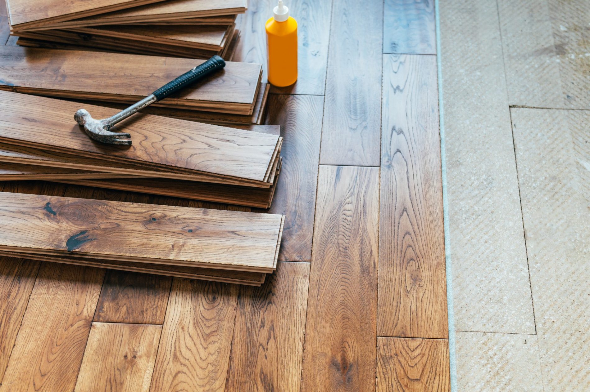 The image displays a wooden floor partially covered by a stack of laminate flooring planks. On top of the planks lies a hammer that might be used for the installation process. Near the stack, there is a bottle, likely containing adhesive or some other installation aid. It appears that a flooring installation or renovation project is underway. The transition between the existing floor and the bare subfloor can be seen to the right in the image.