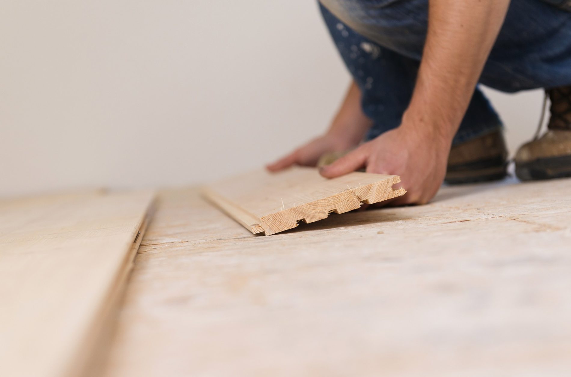 The image shows a close-up of a person's hands and knees installing a piece of tongue and groove wood flooring. The individual is likely kneeling on an unfinished floor and is positioning a wooden plank into place. The flooring piece has a distinctive profile that allows the boards to fit together snugly. The person is wearing casual clothing, including denim and sturdy work boots, indicating that they are engaged in a hands-on DIY or construction task. The background is relatively plain, which keeps the focus on the flooring installation process.