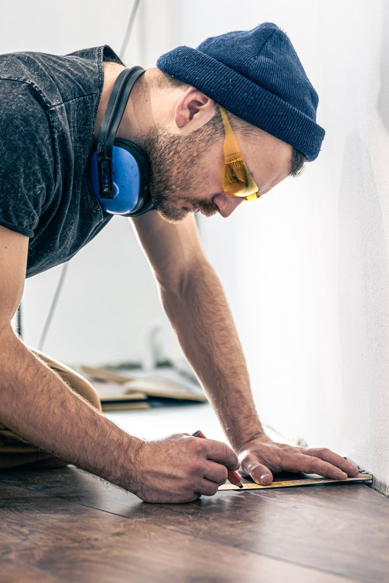 The image shows a person engaged in a carpentry or flooring project. The individual is wearing safety equipment such as ear protection and yellow tinted safety glasses. They are also wearing a knitted beanie. The person appears to be concentrating on measuring or marking a piece of wood flooring with a pencil and a metal ruler, possibly ensuring a precise cut or fit. The setting suggests an indoor environment under renovation or construction where flooring is being installed or repaired.