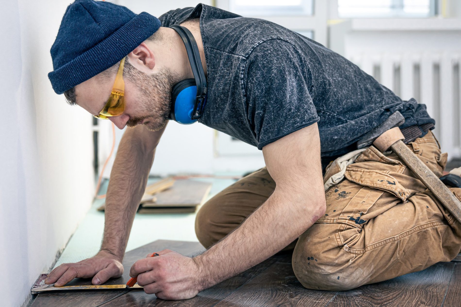 The image shows a person engaged in some sort of construction or renovation work. The individual is kneeling down on the floor, wearing a beanie cap, safety goggles, and blue ear protection headphones. They are dressed in a casual, work-appropriate manner with a dark grey tee shirt and heavy-duty brown pants that have pockets suggesting they might be a type of carpenter or utility pants. This person is measuring and marking a piece of material, possibly to cut or install it as part of the flooring. There are additional materials and tools around, indicating an ongoing project. The setting appears to be indoors, with a white wall and a radiator in the background, which may imply a residential environment. The person shows a focused demeanor, indicating careful attention to the task at hand.