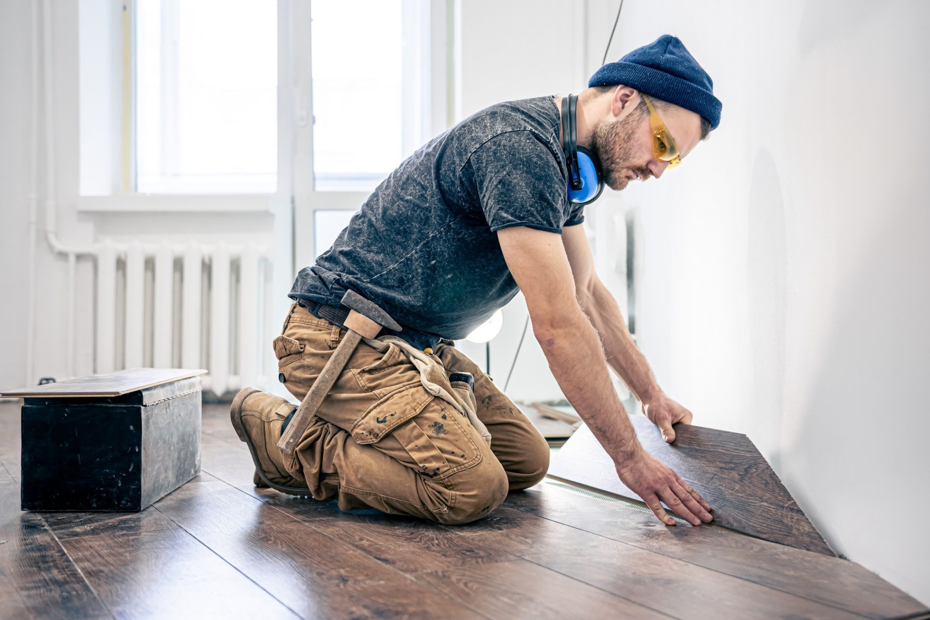 The image shows a person in the process of laying a laminate wood floor. The individual is wearing casual work attire, including brown work trousers, a dark t-shirt, a beanie, and yellow protective eyewear. They also have a pair of blue earmuffs around their neck, presumably for noise protection. The person is kneeling and precisely fitting a piece of laminate flooring into place next to a previously laid section.In the background, there is a white wall with a radiator attached and a large window allowing natural light into the room. To the left of the person, there is a closed metal toolbox on the wooden floor, indicating that this is a work environment where various tools are likely being used for the flooring installation. The flooring appears to be a darker shade of wood, providing a contrast against the bright and minimalistic room.