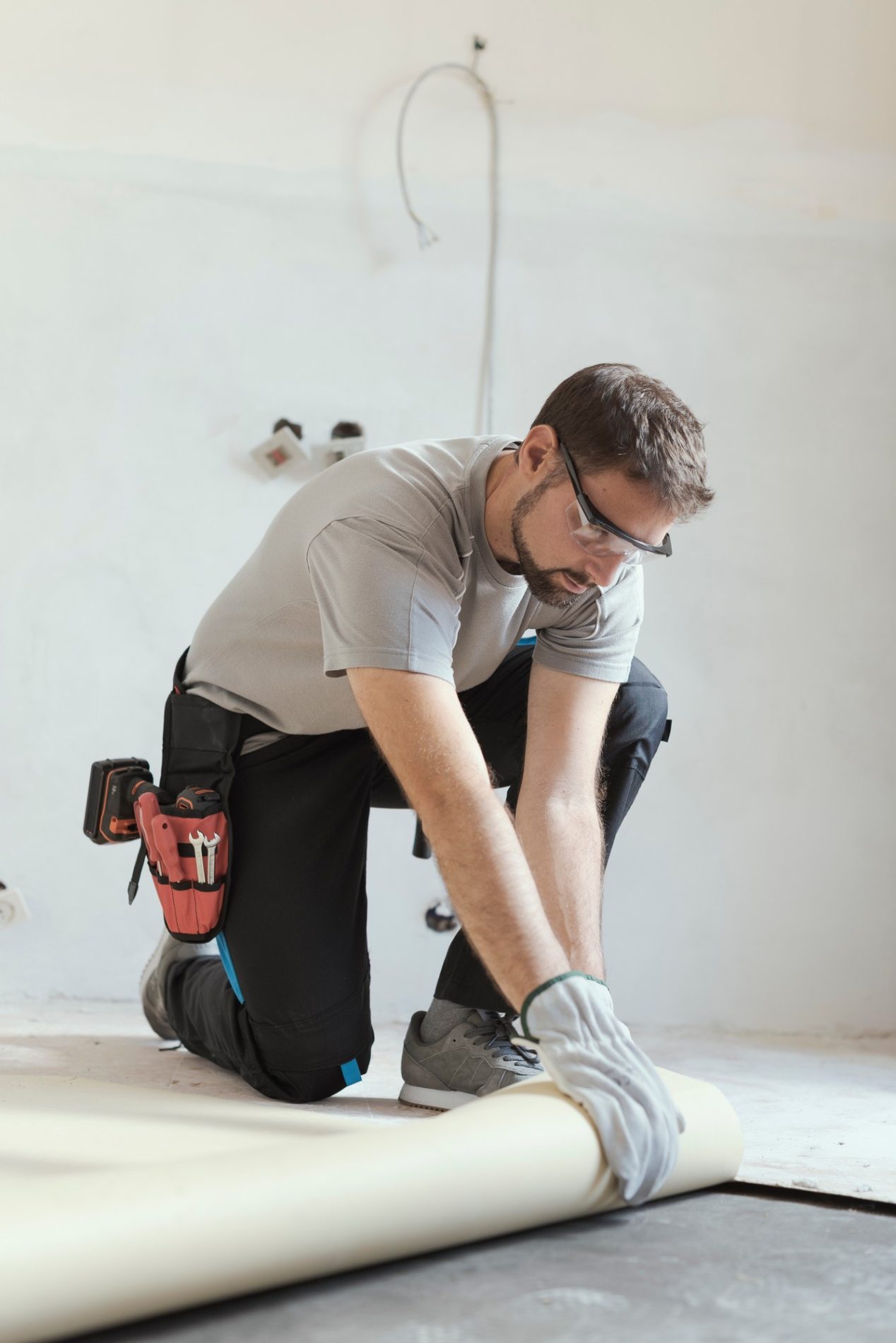 The image shows a person working on a floor. The individual is wearing a gray shirt, black pants with a blue stripe, safety glasses, and gloves. The person has a tool belt with different tools and is in the process of unrolling a sheet of material, likely flooring, on the ground. The walls in the background appear to be unfinished or in the midst of being prepped for painting or other work, suggesting that this might be a construction or renovation site. There are also some electrical wires and outlets visible on the wall in the background.