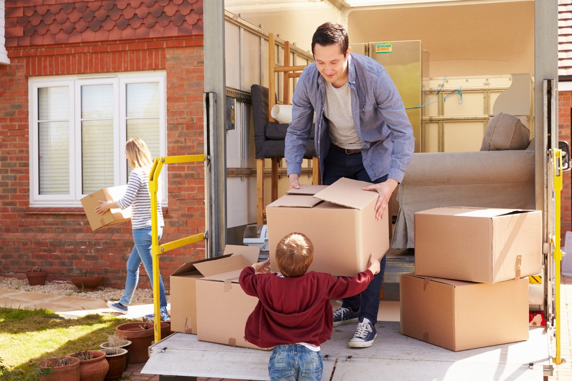 The image shows a family involved in a moving process. A man is lifting a large cardboard box while a child is enthusiastically reaching out to help him with another box. Another individual, who appears to be a woman, is walking away carrying a box and seems to be placing it inside a house. There is a moving lorry open at the rear with a few items, including a sofa, left to be moved. The environment suggests a sunny day in a residential area with a brick house in the background. The scene conveys a sense of cooperation and family activity during the process of relocation.