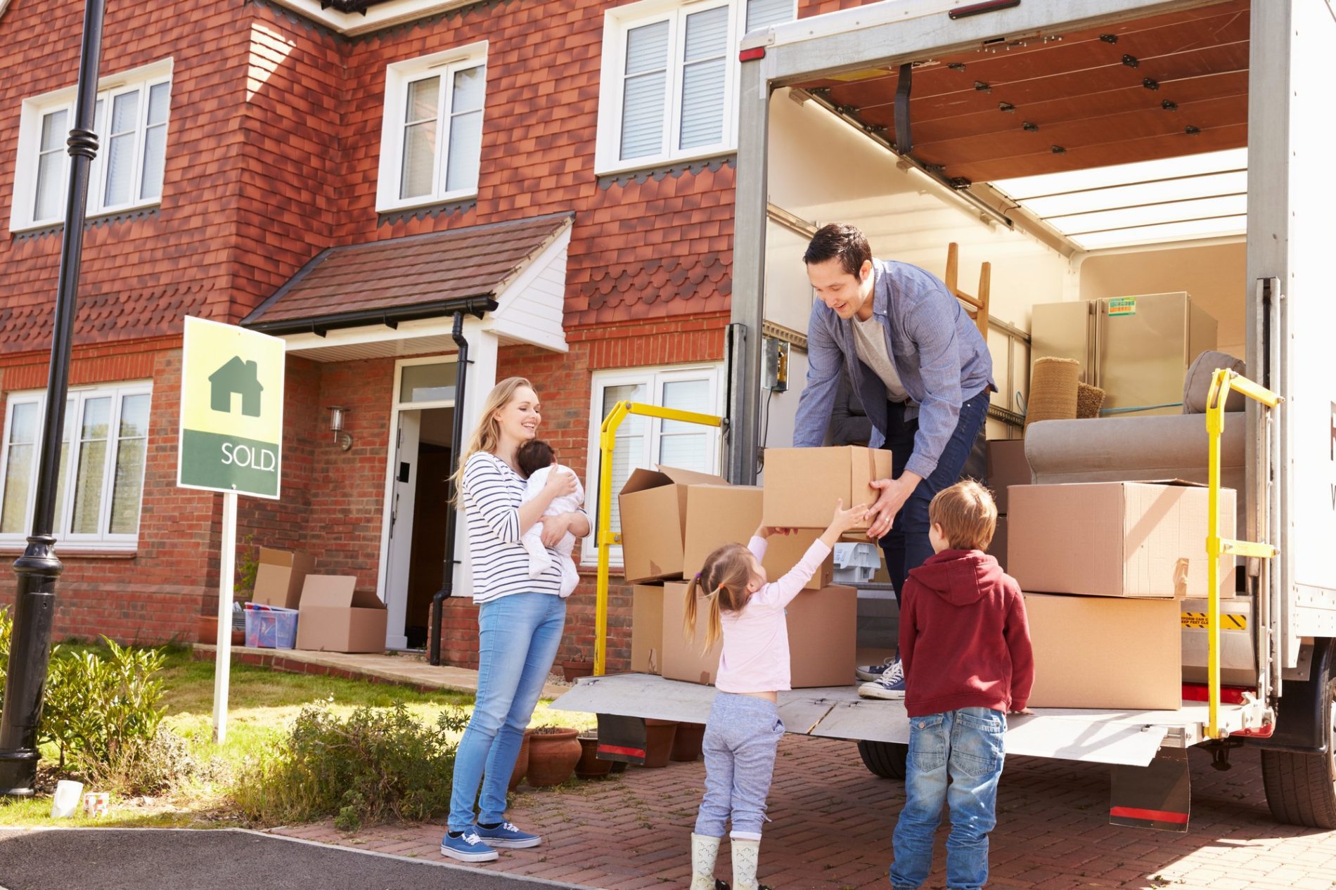 The image shows a family involved in a moving process. You can see a residential brick house in the background with a "Sold" real estate sign posted in the front yard, indicating the house has recently been purchased. A woman is holding a baby and smiling, standing in front of the house, while a man is handing a cardboard box to a young girl. Another child, a boy, is standing next to the girl, looking on. They are beside an open moving van which is loaded with household items, such as boxes and furniture. The scene is set during the daytime under clear blue skies, and the family seems to be in a cheerful mood, suggesting a positive new beginning in their new home.