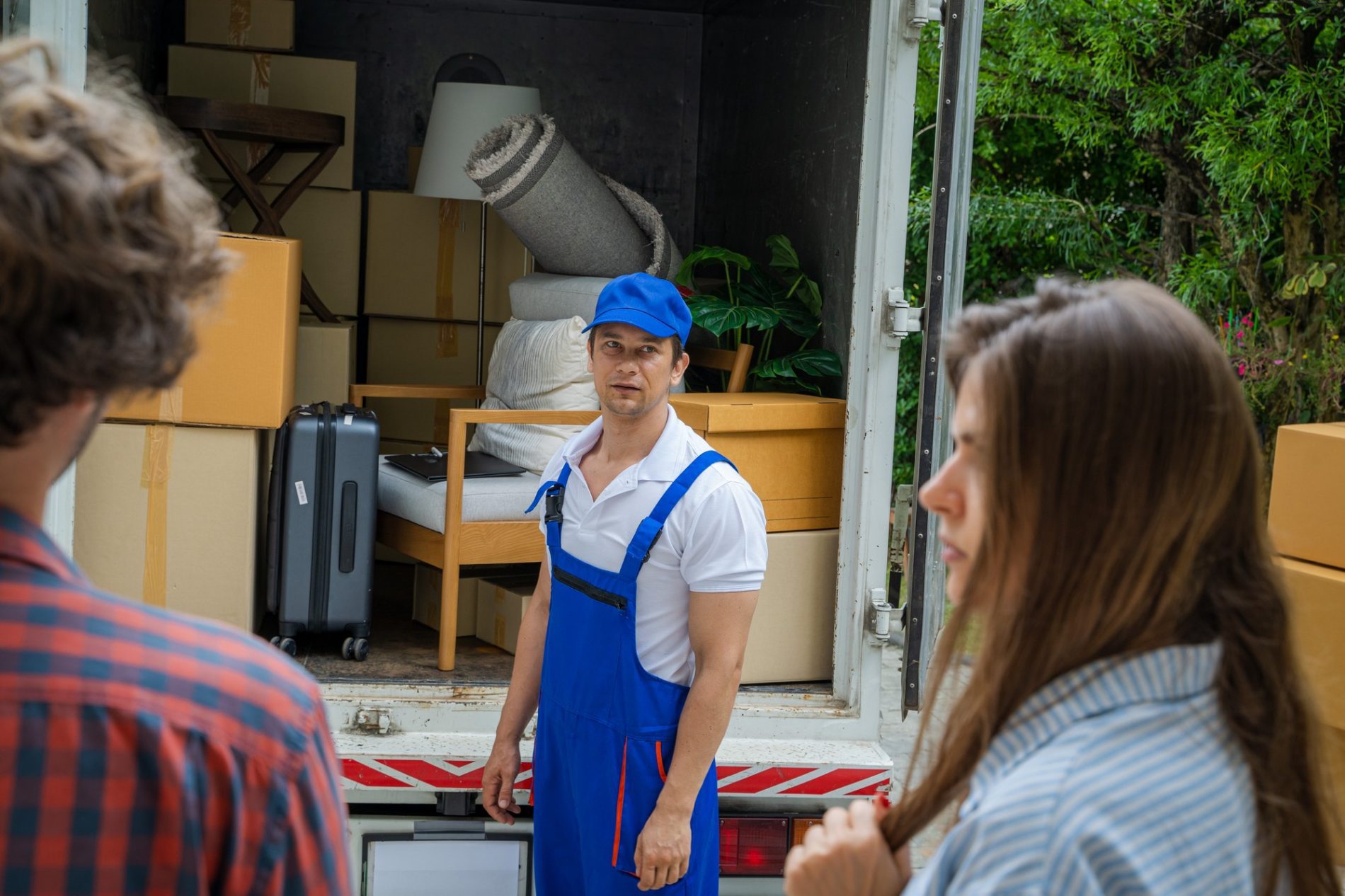 This image shows a moving day scenario. A man in a blue cap and blue overalls, likely a mover, is standing in front of an open moving lorry that's loaded with various packed items such as cardboard boxes, a suitcase, a rolled-up carpet, and pieces of furniture. In the foreground, out of focus, there are two individuals who appear to be the customers, one with the back of his head facing the camera and another looking towards the mover. The setting suggests they are possibly discussing the moving process or preparing to load or unload items. The surroundings have greenery, indicating a residential area.