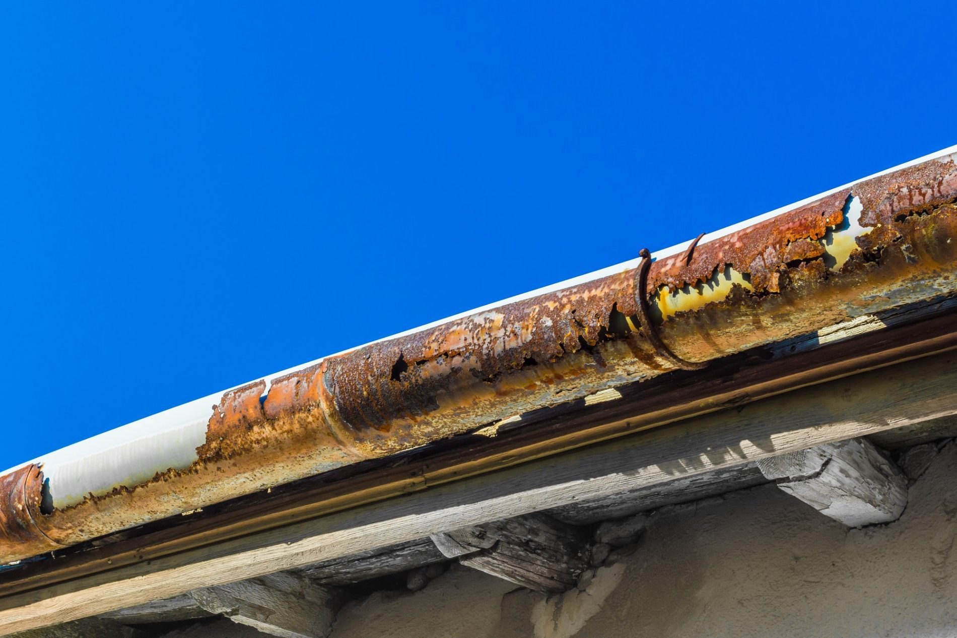 This image shows a close-up view of a building's eave with a heavily rusted gutter. The gutter is corroded, with visible holes and flaking material. Underneath the gutter, wooden eave supports are seen, and they exhibit signs of weathering. The bright blue sky in the background creates a stark contrast with the deteriorating condition of the gutter, emphasizing the need for maintenance or replacement.