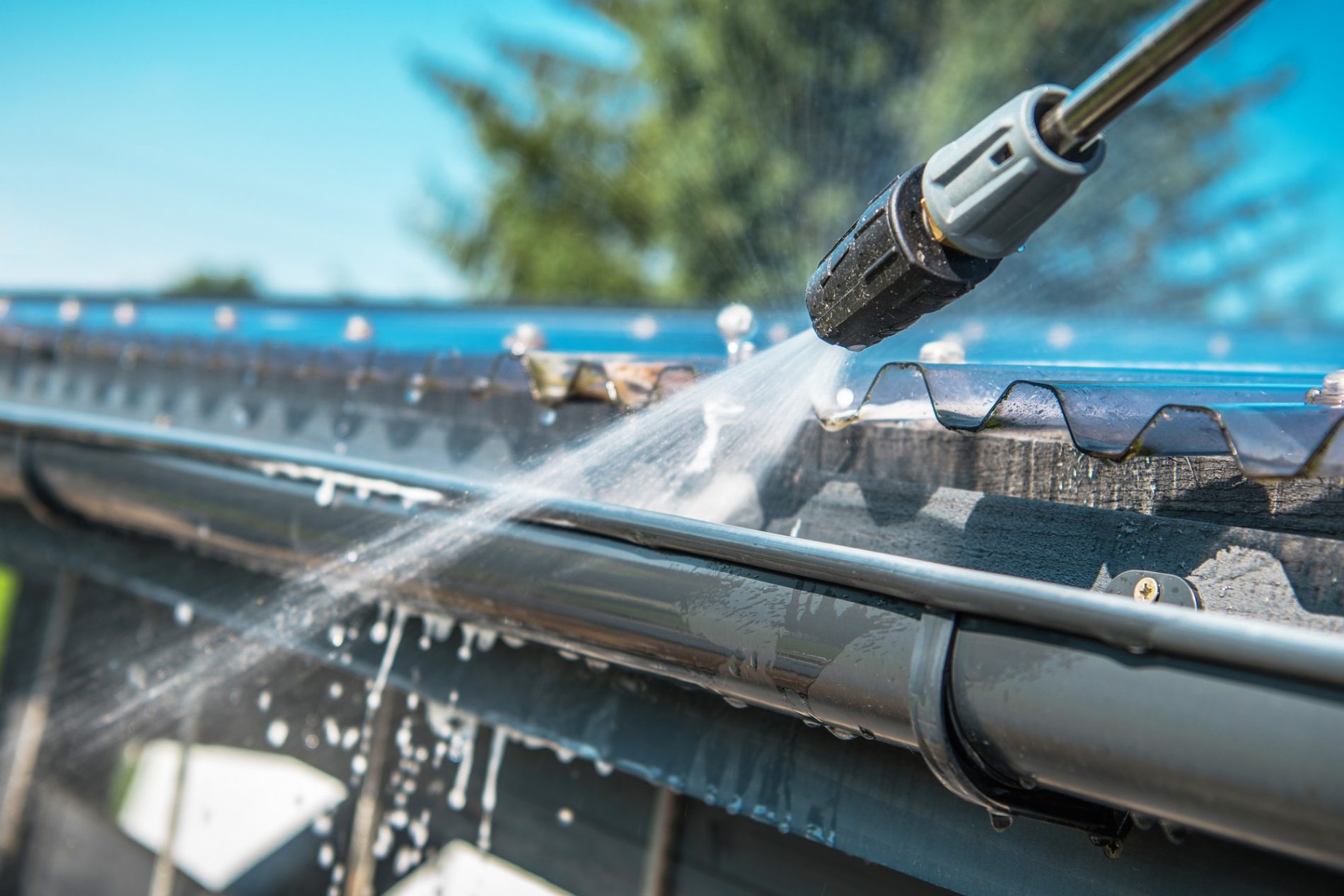 The image shows a close-up of a power washing process. A high-pressure water spray is directed at what appears to be a section of a corrugated metal roof or siding that is attached to a gutter. The water jet is coming from a nozzle attached to a wand or lance, which is likely connected to a pressure washer. The force of the water is visibly removing dirt and probably algae or moss from the surface, and you can see the clean streaks where the water has already passed. This is a common maintenance technique used to clean buildings, roofs, and gutter systems. The background is slightly blurred, suggesting a focus on the cleaning process, and it features a bright blue sky and some greenery, indicating a sunny day, likely in an outdoor environment.