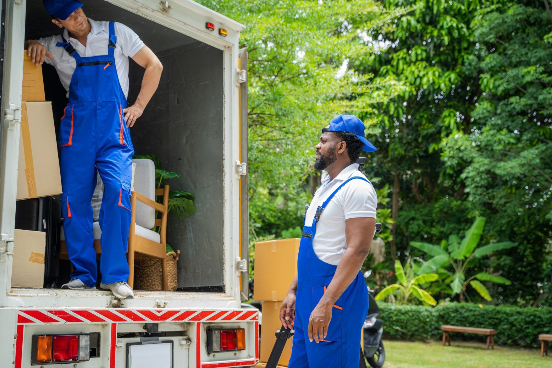 The image shows two people who appear to be professional movers at work. The individual on the left is standing inside the back of a moving lorry, which is open and loaded with various items like cardboard boxes and a piece of furniture that resembles a chair. The man is wearing blue overalls with orange trim and a white t-shirt, along with a cap, and is stepping out of the lorry.The person on the right is standing outside the lorry on the ground, also wearing a uniform similar to the first individual, with blue overalls, a cap, and a white t-shirt. He appears to be holding a moving strap or some tool related to the moving process.The setting is outdoors with greenery in the background, indicating they might be in a residential area. The overall scene suggests that these individuals are in the process of either loading or unloading household belongings as part of a moving service.