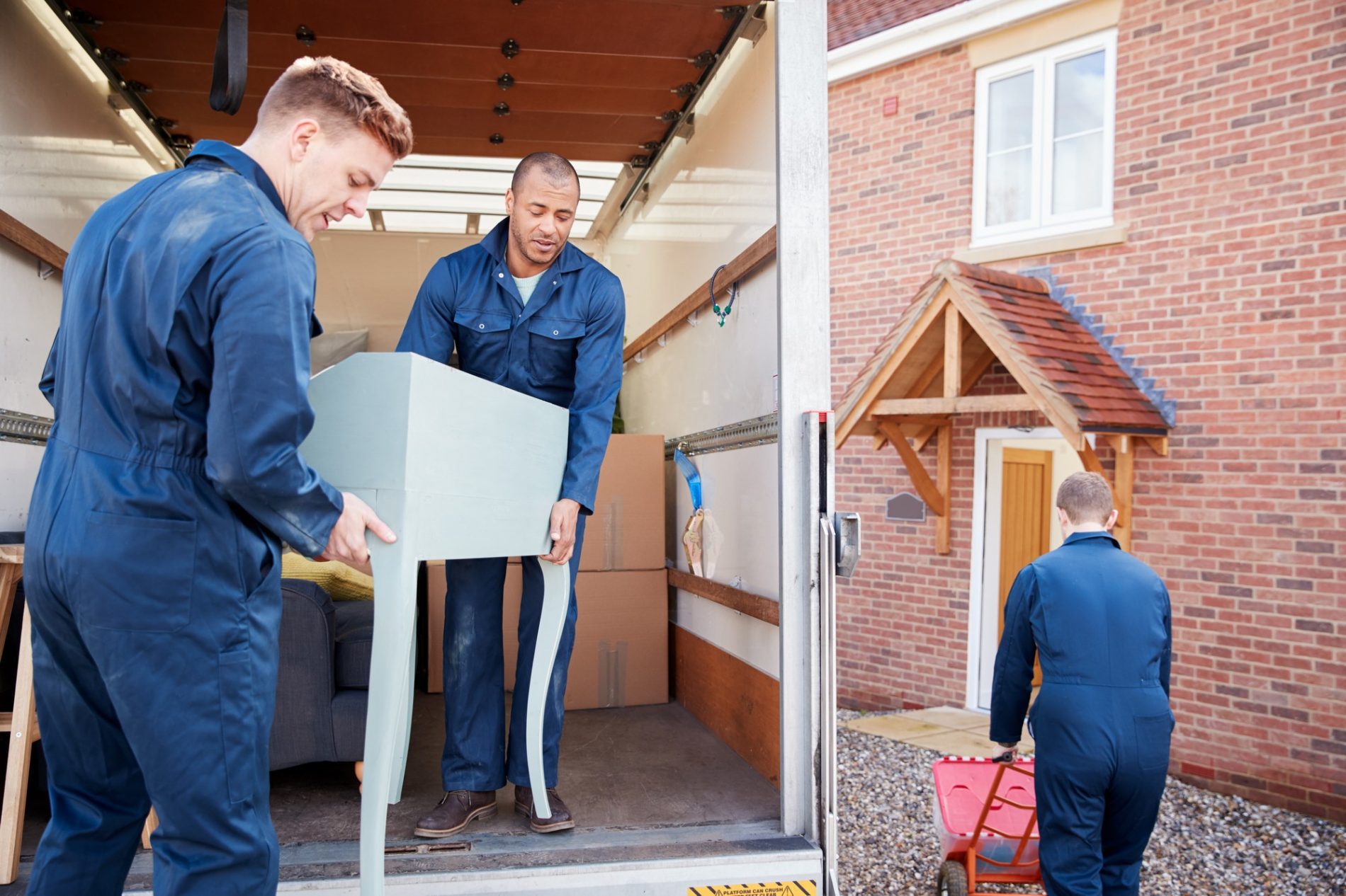 The image shows three individuals who appear to be moving items from a lorry into a residential building. Two of the individuals are carrying a piece of furniture, which looks like a table, from the back of an open moving lorry. The third individual is in the foreground, pushing a red hand lorry or dolly, likely used to help transport heavy items.They are all dressed in blue work uniforms, suggesting they are professional movers. The scene takes place outside of a brick house with a distinctive doorway that includes a wooden arch and a small red-tiled overhang. There are also moving boxes visible inside the moving lorry, indicating an active process of moving items in or out of the house.