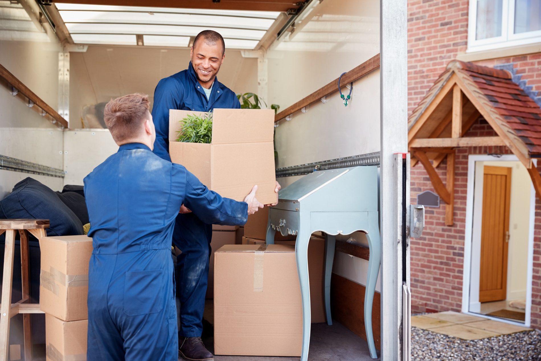 The image shows two individuals engaged in the process of moving. One of them is inside a moving lorry and handing a cardboard box to the other, who appears to be receiving it while standing outside the lorry. They are both wearing matching blue uniforms, which suggests they may be professional movers. The lorry is parked in a residential driveway, and behind them, you can see a house with a brick facade and a wooden door. Inside the lorry, there are several cardboard boxes and a green plant peeking out from one of the boxes, indicating household items being moved. Additionally, there's a piece of furniture, specifically a vintage-style writing desk, also visible inside the lorry. The two individuals seem to be cooperating smoothly and sharing a pleasant interaction as indicated by the smile on the face of the man inside the lorry.