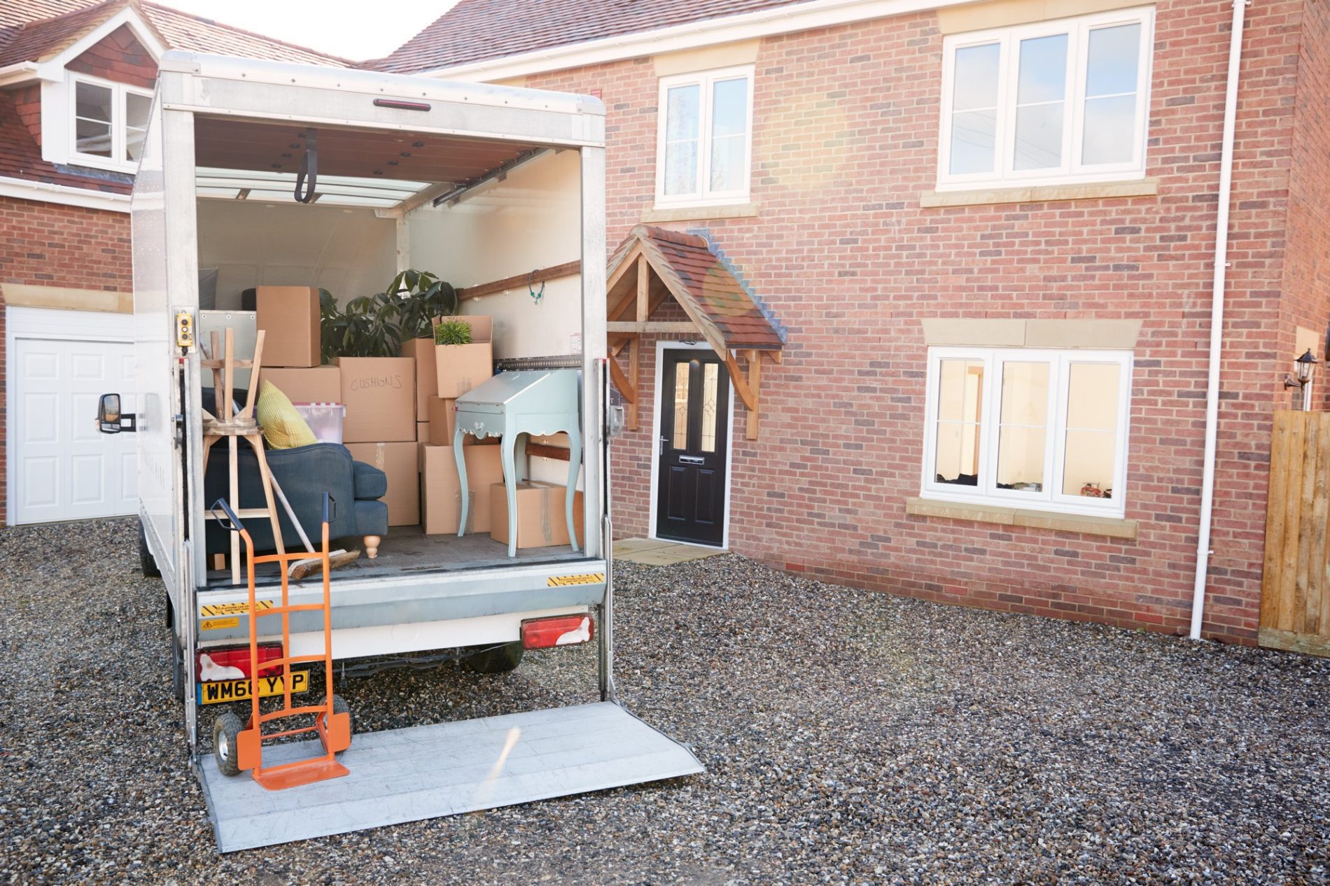 The image shows a moving scene where household items are being loaded into or unloaded from a removal lorry. The back of the lorry is open revealing various items such as cardboard boxes, a couch, and a turquoise-coloured dresser or dressing table. There is a hand lorry (dolly) positioned on the tail lift of the lorry, suggesting items are being moved in or out of the property. In the background, there's a brick house with a white front door and windows, and the driveway is covered with gravel. The setting suggests someone may be either moving into a new home or vacating their previous residence.