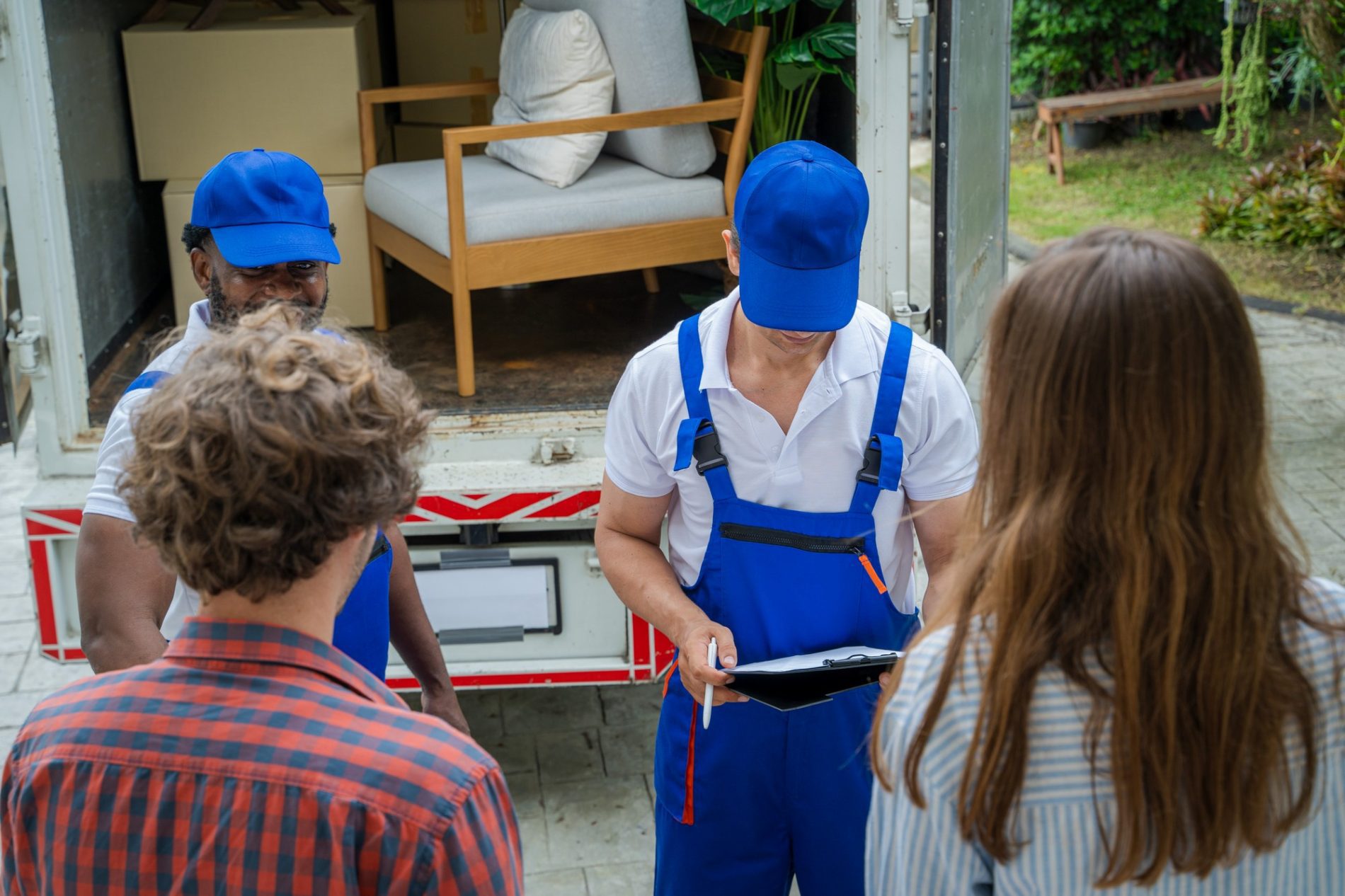 This image features a moving scene where two individuals are presumably the customers, standing and facing a moving company worker who is wearing a blue cap and a white shirt with blue overalls. The worker is holding a clipboard and it appears they are discussing the details of the move or reviewing some paperwork. In the background, we can see an open moving lorry loaded with furniture and boxes, suggesting that the moving process is either about to start or is in progress. They are outside, and a bit of a garden or yard is visible in the far background, indicating a residential setting.