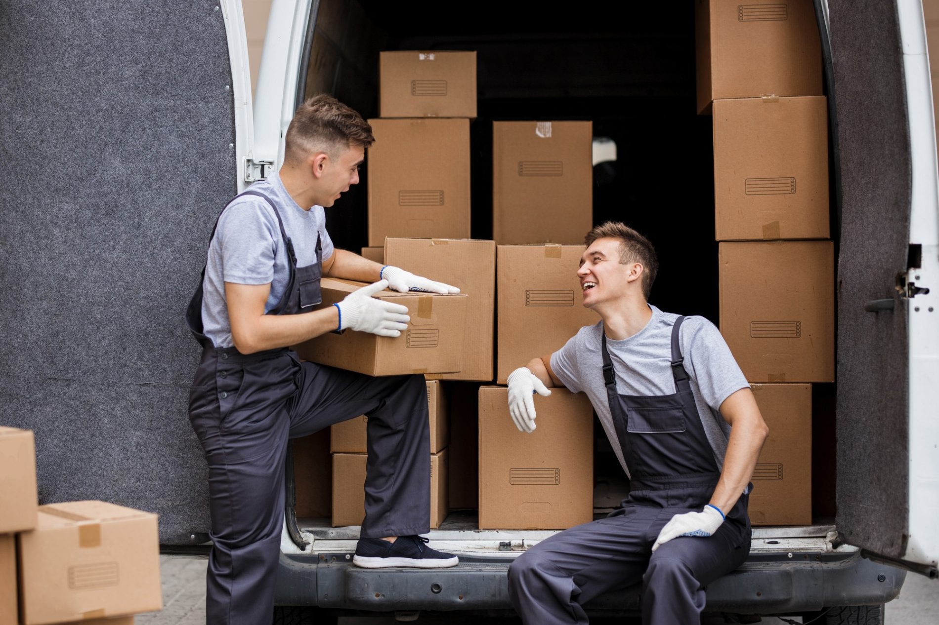 The image shows two individuals wearing work uniforms, engaged in handling cardboard boxes. They appear to be either loading or unloading these boxes from the back of a delivery van. The person on the right is sitting inside the van and is receiving or passing a box to the other person standing outside the vehicle. Both of them are wearing gloves, possibly to protect their hands while handling the boxes. They seem to be in a good mood, sharing a light moment or conversation, as indicated by their smiles and the engaging eye contact. The image reflects a work scenario, likely related to a moving or delivery service.