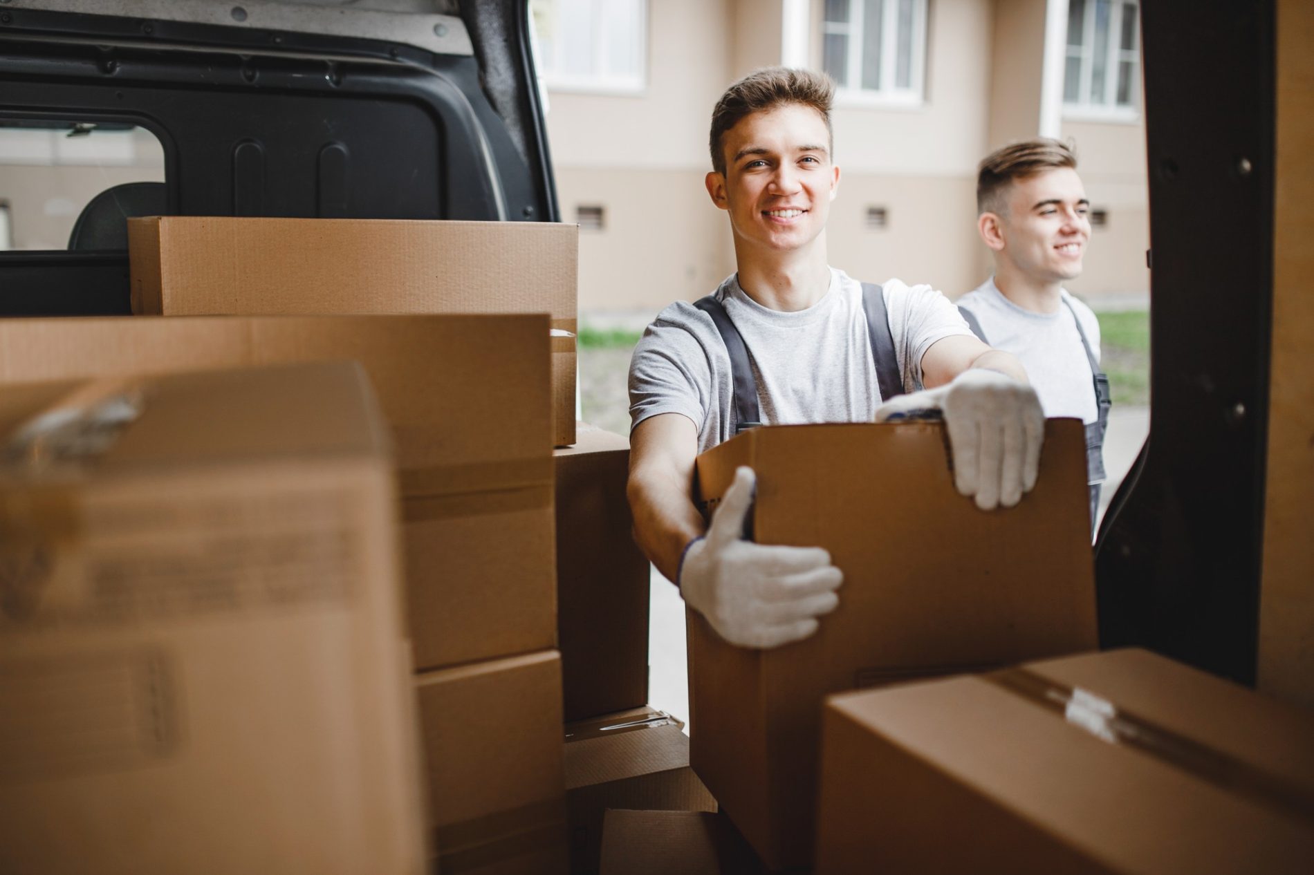 This is an image of two young men who appear to be in the process of moving boxes. They are both wearing casual clothing and gloves, which suggests they are handling the boxes to protect their hands. The boxes are cardboard, and one of the men is lifting a box while smiling at the camera. The open back of a vehicle, most likely a van or large SUV, is visible, and there are multiple boxes inside it, suggesting that these individuals are either loading or unloading the vehicle as part of a move or delivery. The location looks like a residential area, and a building with windows can be seen in the background. There is a sense of activity and positivity conveyed by the expressions of the people in the image.