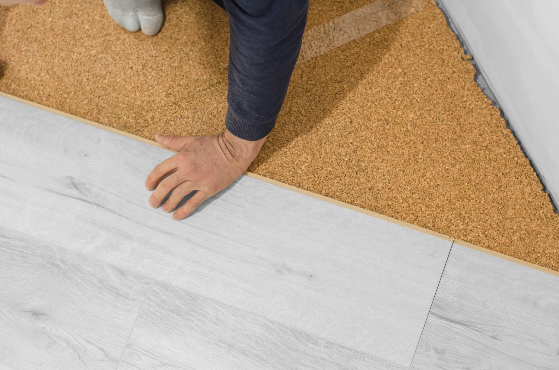 The image shows a close-up of a floor at the threshold of a room, where two different types of flooring materials meet. On one side, there's a light-coloured laminate or vinyl plank flooring with a wood grain texture, and on the other side, there's a tan cork flooring. There's a person's hand touching the cork floor, and the feet of another person are partly visible; one foot is on the cork flooring while the other is on a softer surface like a carpet or a mat, which appears to be gray. The wall adjacent to the cork flooring has a white baseboard.