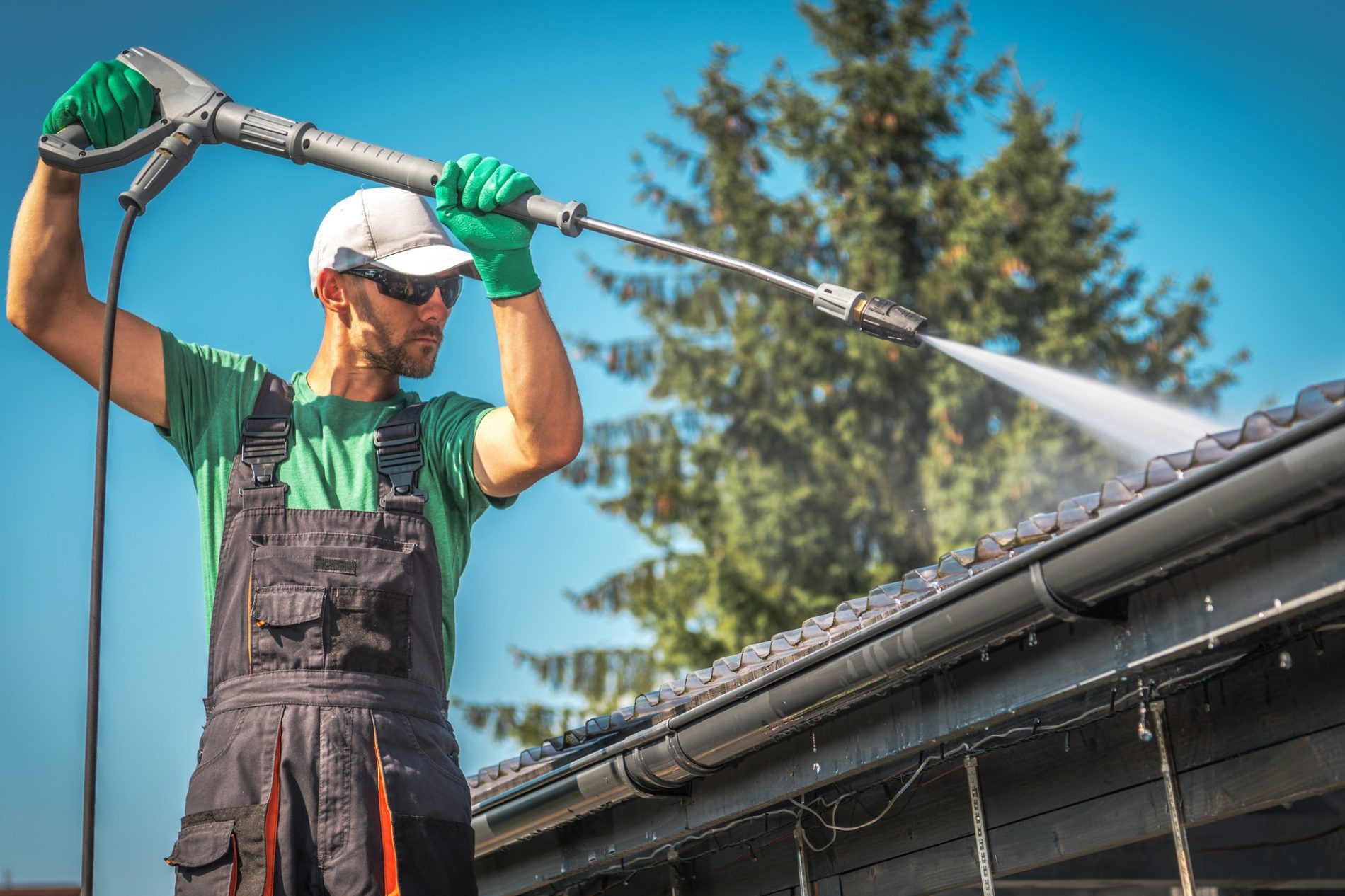 The image features a person cleaning a house's gutter with a high-pressure water cleaner. The person is wearing protective gloves, a white cap, sunglasses, and a work outfit with overalls. They are standing on a ladder and directing the water jet onto the roof gutter to clear away any debris or buildup. The background shows clear blue skies and some trees, giving the impression of a sunny day, which is appropriate for outdoor work like this. The scene portrays a regular maintenance activity typically done to ensure proper water drainage from roofs.