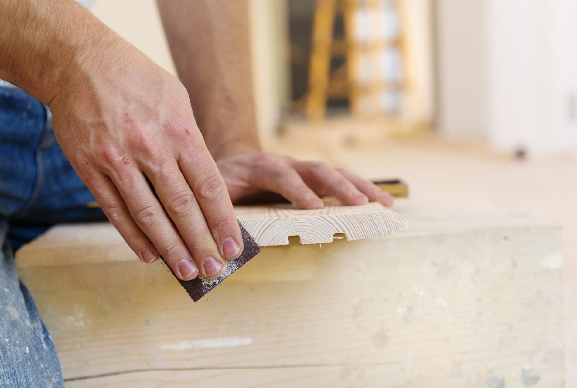 The image shows a person's hands using a piece of sandpaper to smooth or sand a wooden board. The board appears to have a profile or contour cut into it, possibly for decorative molding or trim. The context looks to be a woodworking or carpentry setting. There are wood shavings and dust on the board and hands, indicative of the sanding process, and the person is likely focusing on creating a smooth finish on the wood. The background is blurred, but it seems there is a construction or workshop environment with a ladder faintly visible, giving the impression that this is part of a larger project.