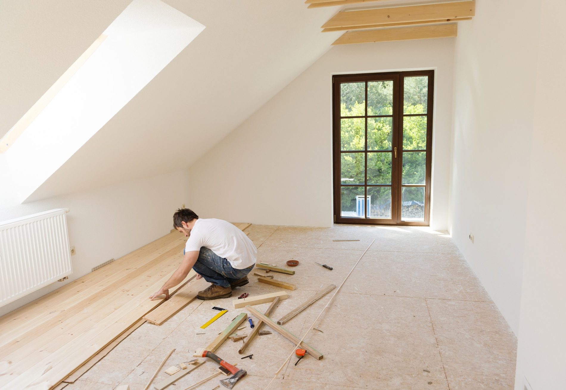 The image shows an interior space undergoing renovation or construction. A person is kneeling on the floor, apparently involved in the process of installing or repairing a wooden floor. There are various tools and pieces of wood scattered around the individual, suggesting active work. The space has sloped walls indicative of an attic or upper story room, with a large window allowing natural light to enter. The room appears partially finished with unpainted drywall, a radiator under the window, and an unfinished wooden floor.