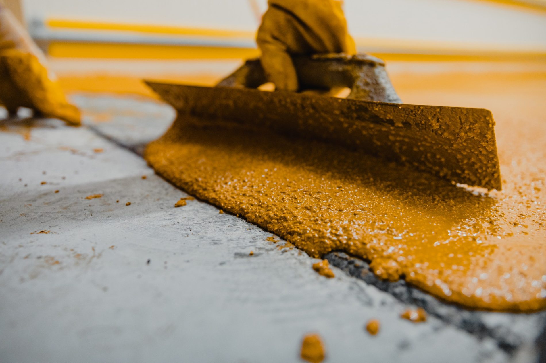 This image shows a close-up view of a person in a yellow protective coverall working with a large flat tool or spreader, which is being used to distribute a thick, golden-brown substance across a flat surface. The substance might be a type of resin, industrial chemical, or perhaps some form of food ingredient. The exact nature of the material isn't clear, but the context suggests some sort of manufacturing, processing, or construction work requiring protective clothing and careful application of the material. There's a strong yellow colour cast in the image, which could be due to the lighting in the environment or due to the colour of the material itself. The focus is on the action being performed and the material, making it an image likely showcasing manual labour or industrial processing.