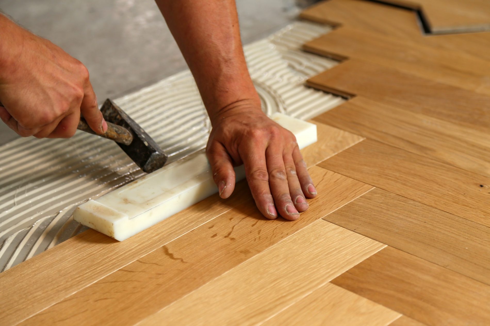 The image shows a person engaged in a flooring installation process. The person's hands are visible – one hand is using a rubber mallet to tap a piece of parquet flooring into place, ensuring a tight fit with the adjacent pieces. Below the flooring is a layer of adhesive or mortar with trowel marks, indicating that the flooring is being glued down to the subfloor. The wooden parquet blocks have a tongue and groove design, which helps in locking the pieces together. The hands are dusty, suggesting active work, and some loose debris can also be seen on the floor, which is common during such installations.