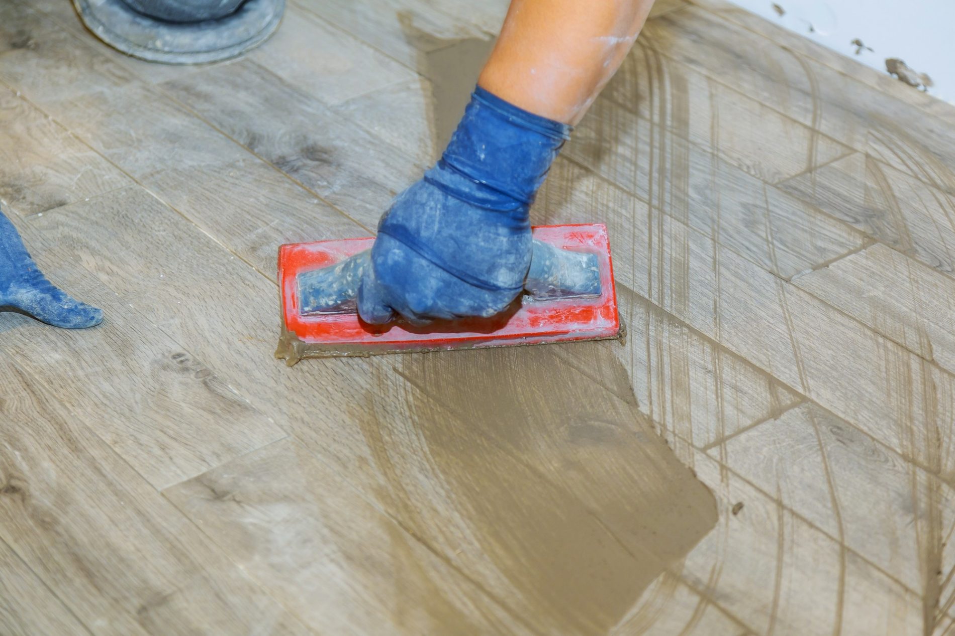 The image shows a close-up view of a person's arm and hand, wearing a blue rubber glove, as they are using a float to apply grout to what appears to be a floor with wood-effect tiling. The floor has visible excess grout on its surface, which is typically spread over and pushed into the gaps between tiles during the process of tiling. This step is important to ensure that the spaces between the tiles are sealed and the finish is smooth. The person seems to be in the process of a home improvement project or some sort of construction work involving tile setting.