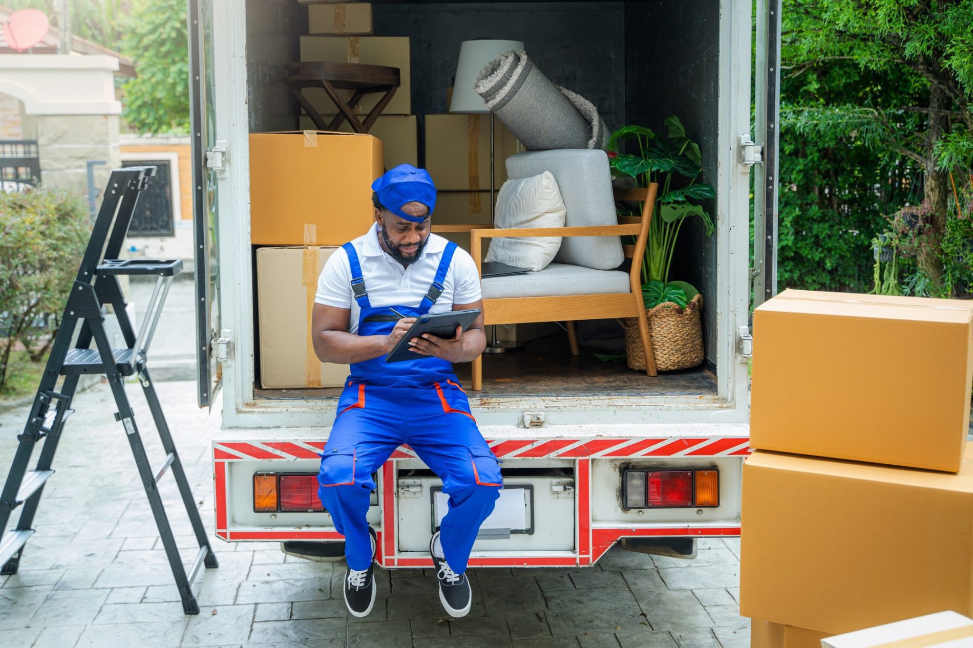 The image depicts a man in a blue and white work uniform, possibly a mover, sitting on the edge of the open back of a moving lorry. He is focused on a digital tablet he's holding. The interior of the lorry is partially visible and contains a variety of items such as boxes, some furniture, a rolled-up rug, and a potted plant. There's also a step ladder placed on the ground to the left, suggesting packing or moving activities. On the right side of the image, there are some cardboard boxes placed on the ground, again indicating a scene of moving out or moving in. The setting appears to be residential, with greenery and part of a building visible in the background.