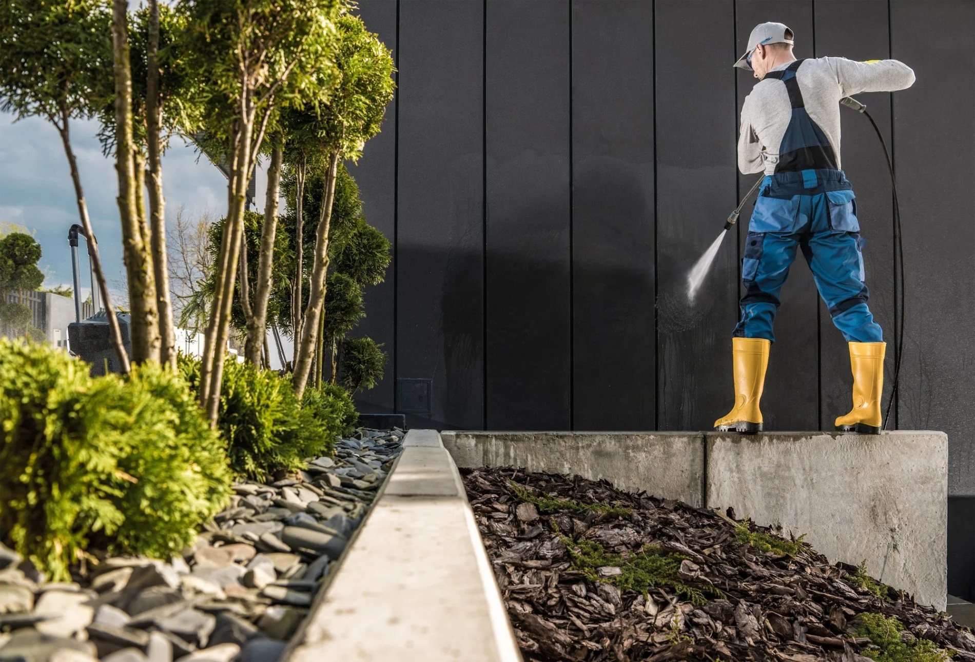 The image shows a person wearing yellow rubber boots, blue waterproof pants with braces, and a grey long-sleeved shirt, using a high-pressure water spray to clean a dark wall. The person is wearing a white cap, suggesting outdoor workwear typical for maintenance or cleaning tasks. The left side of the image features landscaping with small trees, shrubs, and a bed of rocks and mulch. The day seems partly cloudy with sunlight, and the setting looks like a modern, landscaped outdoor area, likely part of a building's exterior environment.