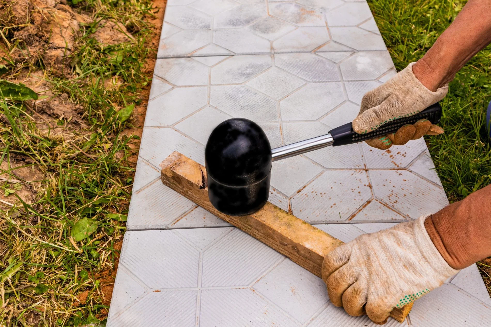 The image shows a pair of hands, one wearing a glove, using a rubber mallet to strike a piece of wood. This is likely a scenario where someone is engaging in a construction or landscaping project involving the laying of tiles or pavers. The wooden board is possibly being used to evenly distribute the force of the rubber mallet to properly set or adjust the position of the tiles. The setting appears to be outdoors, as indicated by the grass and dirt visible at the edge of the tiled area.