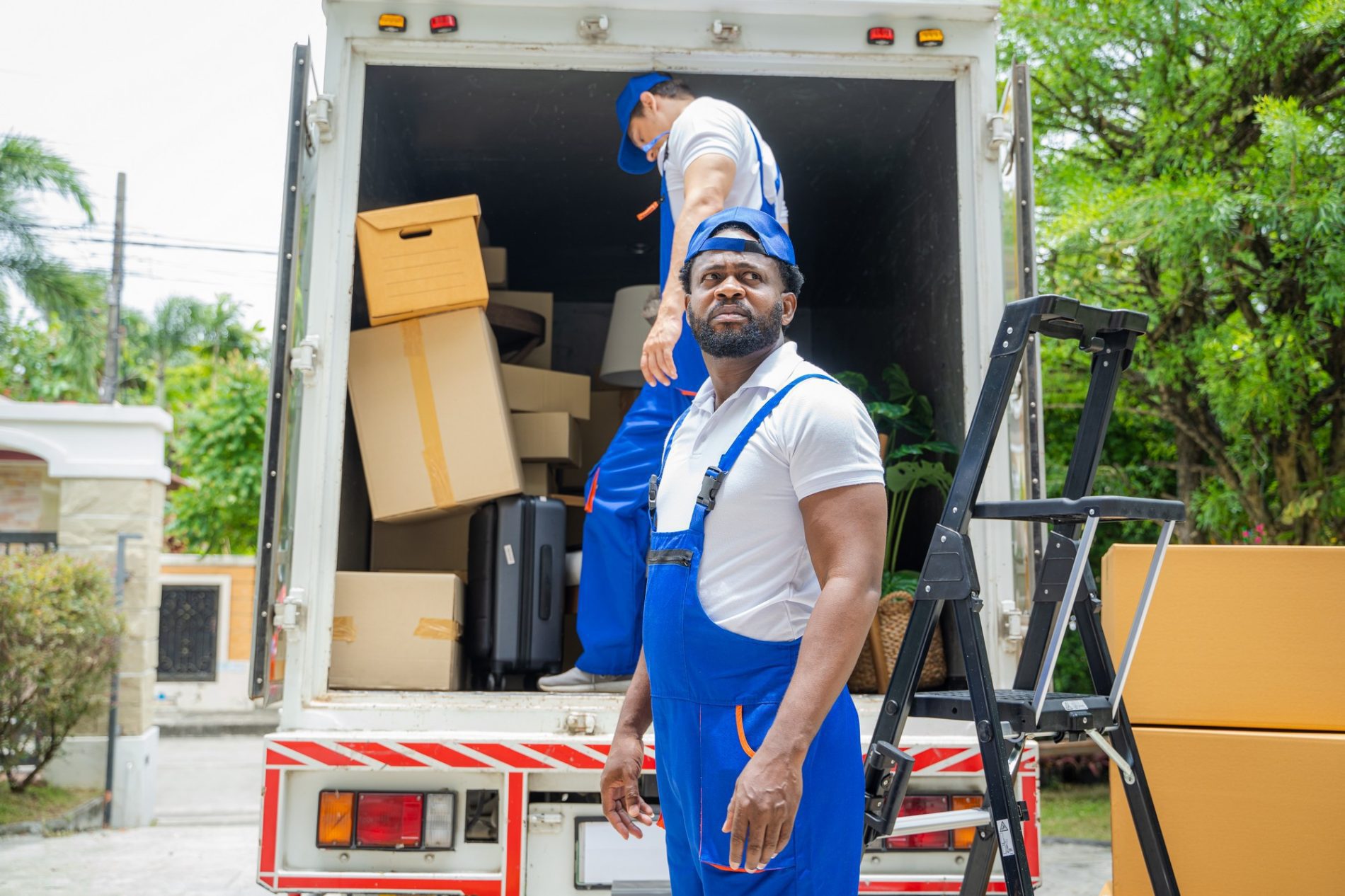 This image shows two people who appear to be moving professionals in the process of loading or unloading household items from a moving lorry. The man in the foreground wears blue overalls with a white shirt and has a confident stance, looking directly at the camera. The second individual is inside the lorry, organizing various items. The moving lorry is partially full of cardboard boxes, some stacked neatly while one appears to be tipping over. There is also a black suitcase and a folded ladder resting against the back of the lorry. The environment suggests the location could be a residential area, as suggested by the presence of a house with a gated fence in the background. The weather looks fair, and it seems to be daytime.