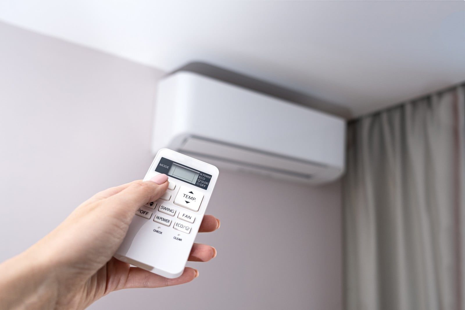 The image shows a person's hand holding a remote control pointed at an air conditioning unit mounted on a wall. The scene indicates that the person is likely adjusting the settings of the air conditioner. The remote has multiple buttons, including ones labeled "MODE," "TEMP," "SWING," "FAN," and "ECO," presumably for different functions of the air conditioning system. The room appears to be an indoor environment with a curtain partly visible on the right side of the frame.