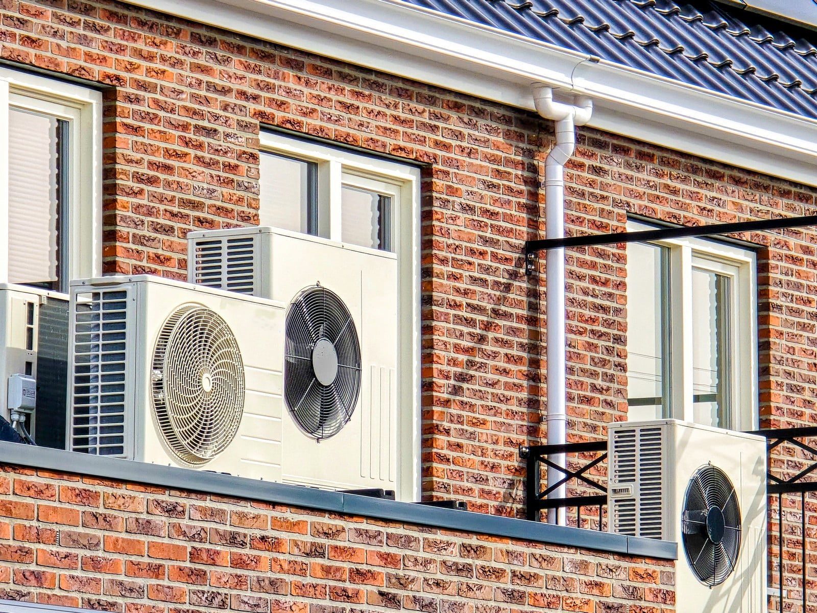 The image shows the exterior wall of a brick building with three outdoor units of split-system air conditioners mounted on it. The wall has a couple of windows with closed blinds. The roof of the building has dark roof tiles, and there is a white drainpipe running vertically down the wall. The air conditioner units are situated on metal brackets that have been secured to the building's structure. The fans are visible on each unit, indicating they are likely in operation or ready for use. The time appears to be during the day due to the bright lighting and shadows on the building.