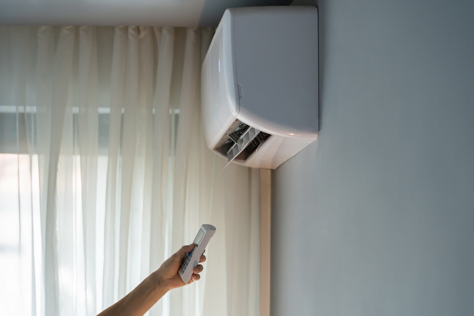 The image shows an interior room where a person is holding a remote control aimed at an air conditioning unit mounted on a wall. The air conditioner appears to be switched on, with its flap open, signalling that it may be actively cooling the room. In the background, semi-transparent curtains cover a window, diffusing the light entering the room. The image captures a moment of interaction between the user and the appliance, possibly to adjust the temperature or settings of the air conditioner.