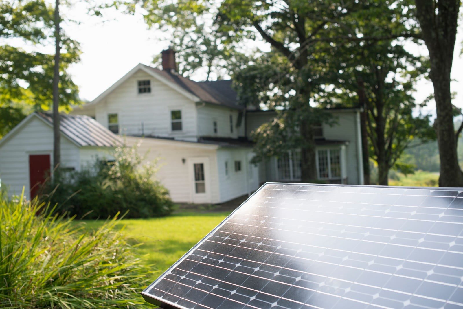 The image features a corner of a solar panel in the foreground, which is sharply in focus. The background, slightly out of focus, shows a white house with greenery and a large tree, indicating a residential setting, possibly suggesting the use of solar energy in a home environment. The sunlit conditions and the presence of the solar panel illustrate the context of renewable energy generation.