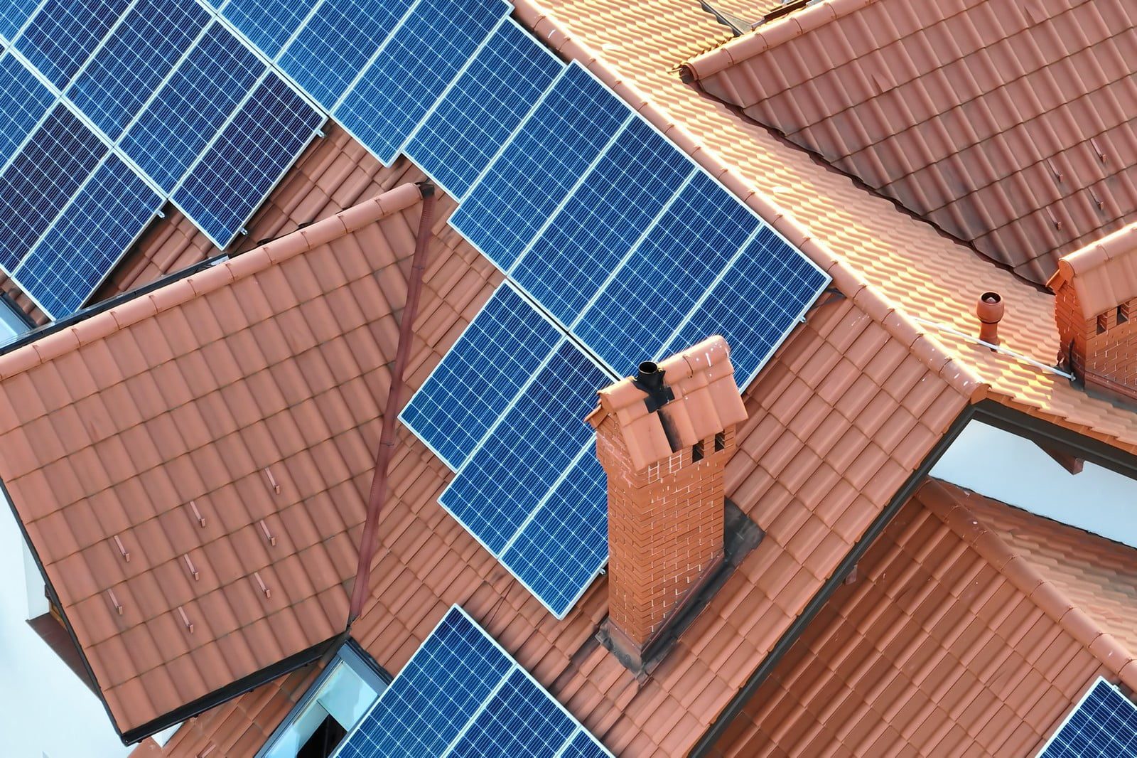 This is an aerial image showing a section of a building's roof with solar panels installed. The solar panels are blue in colour, indicating that they are most likely made of silicon, which is common for photovoltaic cells. The terracotta-coloured roof tiles are typical of residential construction. There are also several brick chimneys visible, which further suggests that this is a residential building. The shadows cast by the chimneys indicate that the sun is shining from the side, and the reflection on one of the panels might suggest it's either early or late in the day. This setup represents a household's investment in renewable energy.