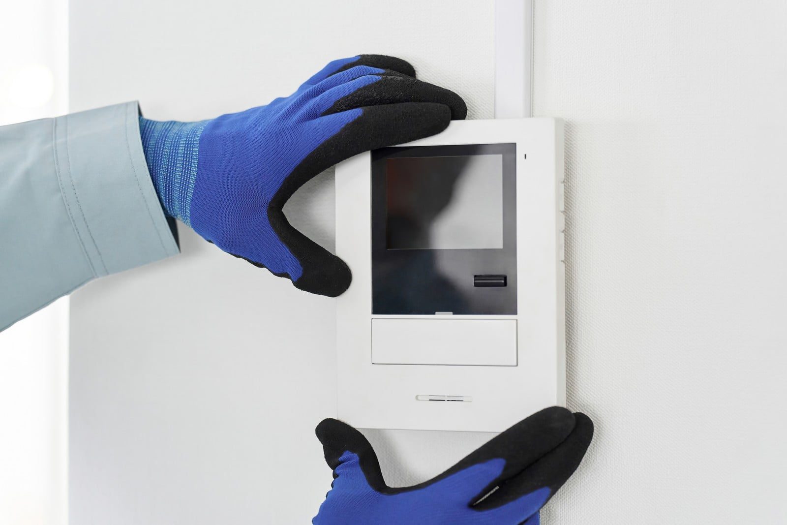 The image shows a pair of hands wearing blue and black work gloves. The hands are interacting with a white wall-mounted device that appears to be an intercom system, access panel, or some sort of electronic control unit. The device has a screen and a few buttons or slots below it, suggesting it could be used to communicate, secure access, or operate a system within a building.