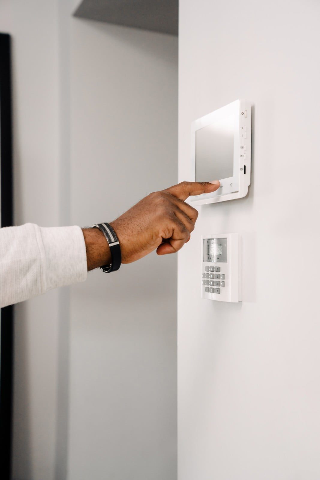 The image shows a person's hand reaching out to interact with a wall-mounted control panel, which likely serves as an interface for a security or home automation system. On the right side of the image, there is a separate keypad security device. Both devices are mounted on a clean, white wall, suggesting a modern and minimalist interior setting. The person appears to be wearing a long-sleeve shirt and a wristwatch. It appears that the individual may be entering a code or setting on the control panel.