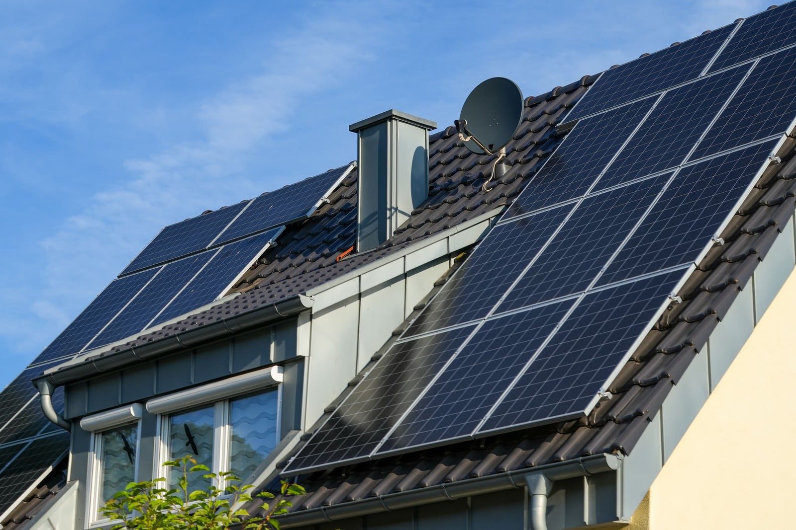 The image shows a residential house with a pitched roof that is fitted with solar panels, which are devices that convert sunlight into electricity. Additionally, there is a satellite dish attached to the roof. The presence of a vent pipe suggests a requirement for ventilation, likely from a bathroom or kitchen. The house appears to have a modern design, as indicated by the window skylights and gutter system for rainwater collection. Overall, this house seems to combine both traditional elements like tiled roofing with modern technology aimed at energy efficiency and functionality.