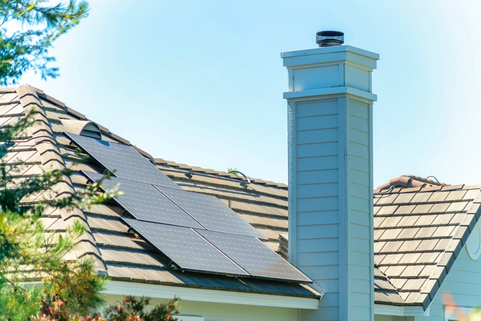 The image shows a house with a sloped roof on which solar panels are installed. There is a chimney extending from the roof with what appears to be a chimney cap on top. The architecture suggests a residential building, and the presence of solar panels indicates a use of renewable energy for electricity generation. The sky is clear, suggesting it's a sunny day, which is ideal for solar energy production. There are also trees partially visible around the structure, providing a touch of greenery.