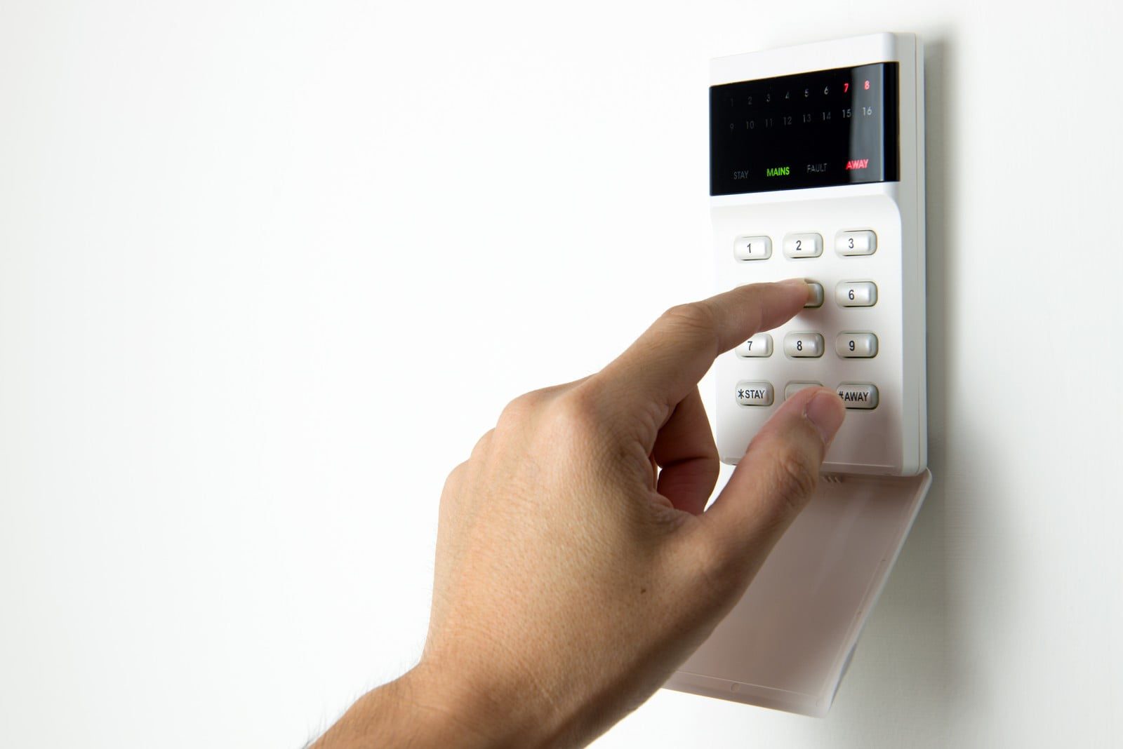 This image shows a person's hand pressing a button on a security alarm keypad. The keypad has a numerical pad for inputting a code, additional function buttons labeled "STAY" and "AWAY", and an LED indicator panel at the top displaying numbers for different zones and status indicators such as "MAINS", "FAULT", and "AWAY". The setting appears to be indoors against a white wall, suggesting this is part of a residential or commercial security system used to arm or disarm an alarm.