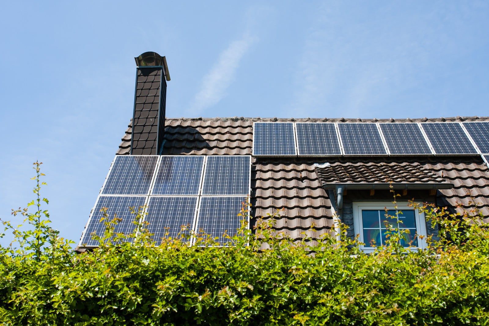 The image displays a section of a house with a pitched roof covered in dark tiles. Mounted on the rooftop, there are several solar panels installed for capturing solar energy. There is a chimney on the left-hand side of the roof. Just below the roof, a dormer window with a sloped roof extension is visible. The bottom of the image shows a lush green hedge, and above, the sky is blue with a few wispy clouds, indicating it might be a sunny day, which is ideal for solar energy generation.