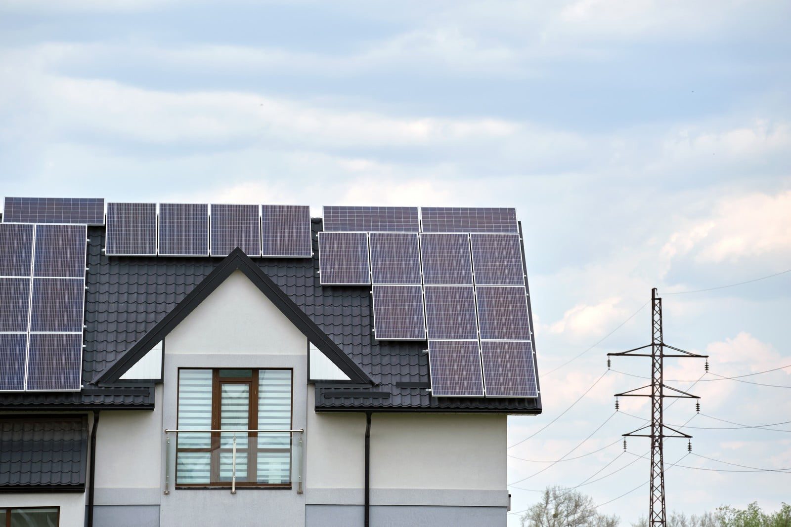 The image shows the upper portion of a modern house with a slanted roof, on which a number of solar panels are installed. These panels are designed to capture sunlight and convert it into electricity. To the right of the house, there is an electricity pylon (also known as a transmission tower) that is part of a network used for distributing electrical power across distances. The sky in the background shows a mix of clouds suggesting it could be either a partly cloudy day or clearing up. The juxtaposition of the house with solar panels and the electricity pylon highlights a contrast between individual renewable energy production and the traditional power grid.