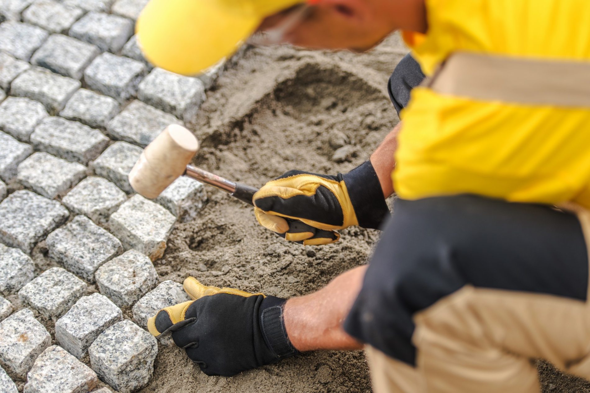 The image shows a person laying cobblestone pavers. The worker, whose face is not visible, is dressed in work gear including a yellow high-visibility shirt, gloves, and what seems to be protective trousers. They are using a mallet to carefully set or adjust a cobblestone into place. The stone is gray and appears to be granite or a similar type of durable material, which is commonly used for paving to create durable and aesthetically pleasing surfaces. The background shows that several pavers have already been laid down. There is also sand visible, which is typically used as a base layer for the pavers to ensure a level and stable surface.