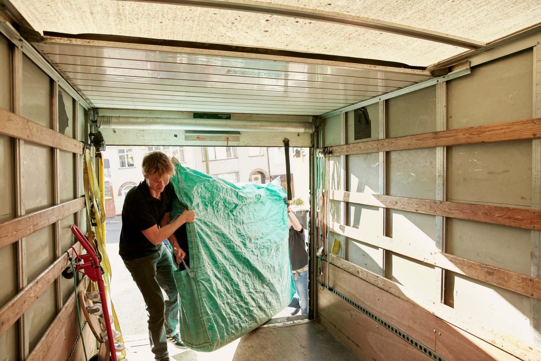 The image shows the interior of a moving lorry or cargo lorry where two people appear to be in the process of moving items. One person is inside the lorry, lifting a large, bulky item that is covered with a green tarp. Another individual who is partially visible seems to be assisting from outside the vehicle. The inside of the lorry has wooden slats on the walls that can be used to secure items during transport, and there's a red dolly near the lorry's opening, implying that it's being used to aid in the moving process. The lighting suggests it is daytime, and the environment outside the lorry looks like an urban area with buildings in the background.