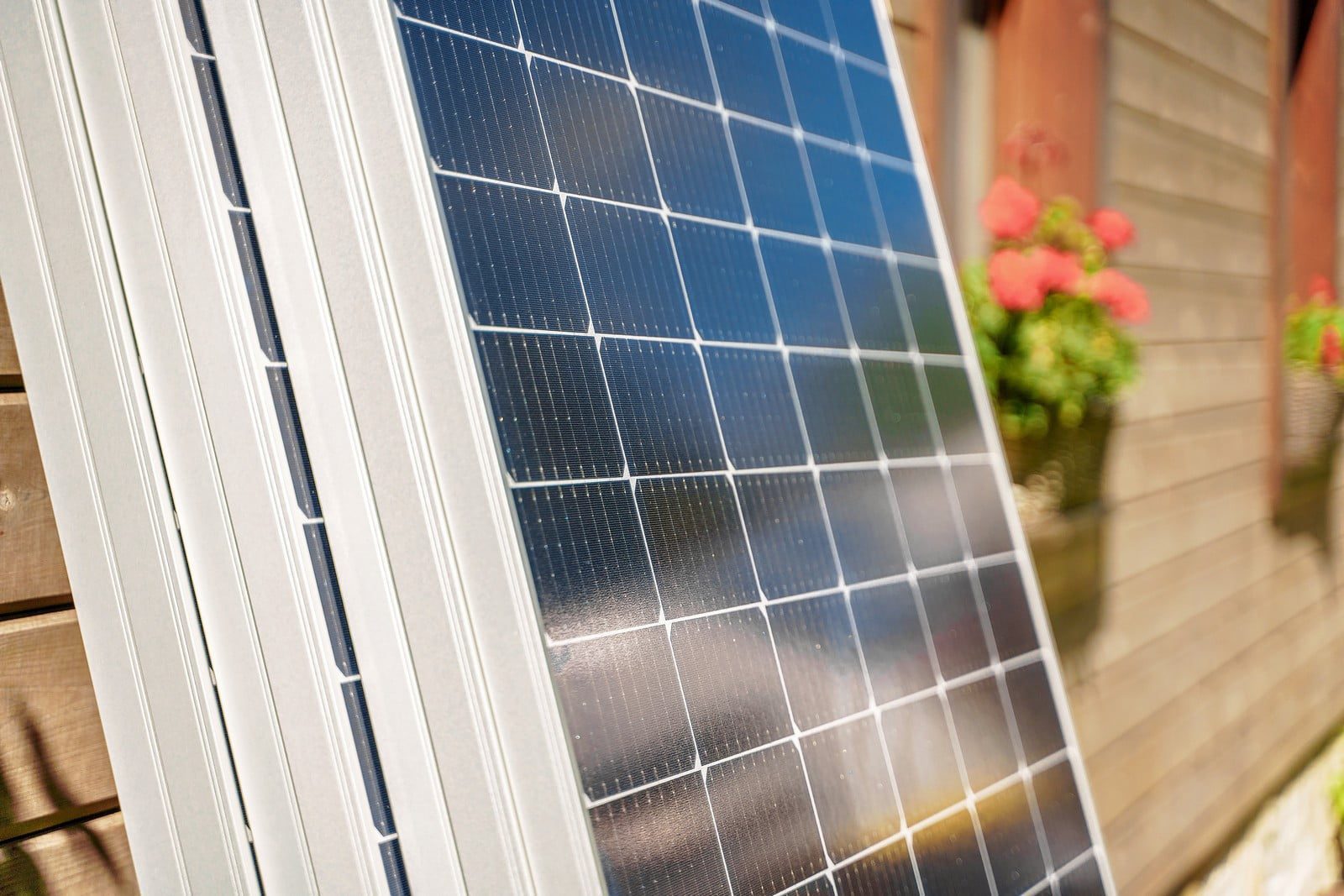 Certainly! Here's a detailed alt text description for the image:"Close-up view of several dark blue solar panels prepared for installation. Each panel is composed of multiple photovoltaic cells aligned in a grid pattern. The cells are encased in a reflective, transparent covering that glistens under natural light. The panels are situated on the ground, indicating they are ready to be lifted and mounted. In the background, there is a blurred view of other installation equipment and possibly some foliage, suggesting an outdoor setting."