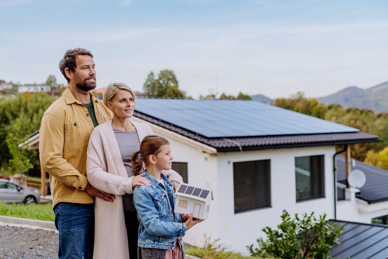 Sure! Below is a detailed alt text description for the provided image URL:"A cheerful family of four is standing outdoors on a bright, sunny day. They are holding a paper model of a house decorated with a solar panel on its roof. The family includes a mother, a father, and their two young children—a boy and a girl. The parents, both casually dressed, are standing behind their children with wide smiles. The boy, wearing a green T-shirt, and the girl, dressed in a pink dress, are holding the model house between them, emphasising their collective enthusiasm for sustainable living. The background features a lush, green landscape with clear blue skies, symbolizing a clean and eco-friendly environment."If there's any more detail or any specific aspect of the image you'd like to emphasize, please let me know!