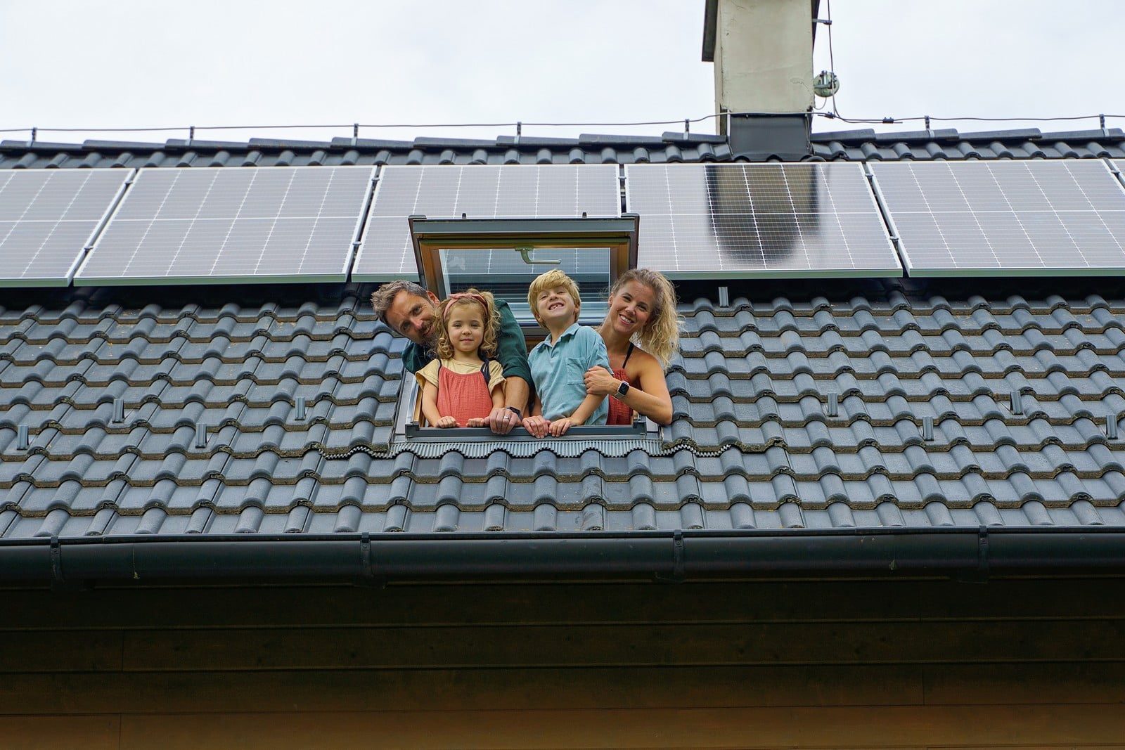 The image depicts a joyous family of four leaning out from a skylight window on a rooftop, against a clear blue sky. Each member of the family appears animated and smiling, embodying happiness and togetherness. The parents, a man and a woman, are positioned slightly behind their two children, a young boy and girl, who are leaning further out of the window and excitedly waving. The man is wearing a casual blue button-up shirt, while the woman is adorned in a light-coloured blouse. The children are both dressed in colorful, playful outfits. The rooftop is covered with traditional clay tiles, and the skylight window is open wide, allowing ample natural light to flood in. The background shows part of the house exterior and a few wispy clouds in the otherwise sunny sky. The overall atmosphere of the image is one of warmth, family bonding, and a cheerful day.